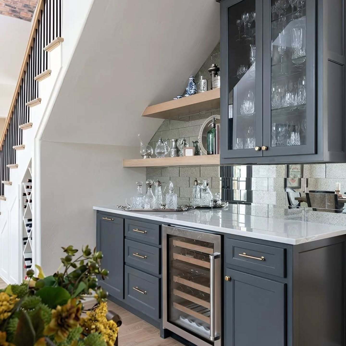 Dark gray custom bar cabinets, glass upper cupboards, and a wine fridge under a staircase.