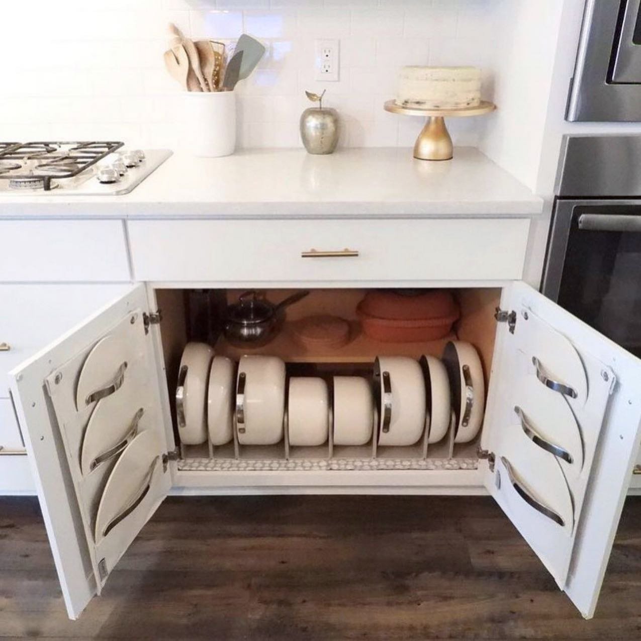 White kitchen cabinet open, showing nesting ceramic pots divided by a wire rack and custom-slotted pot lid storage built into the inner doors.