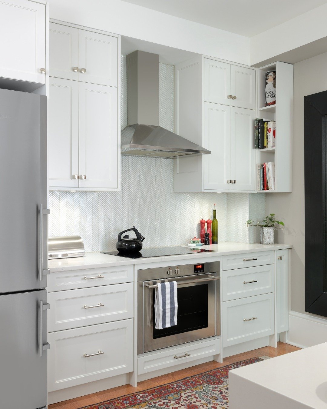 Small, modern white kitchen with white shaker cabinets, a subtle white slim-tile chevron backsplash, a stainless steel oven and hood, and an induction cooktop.