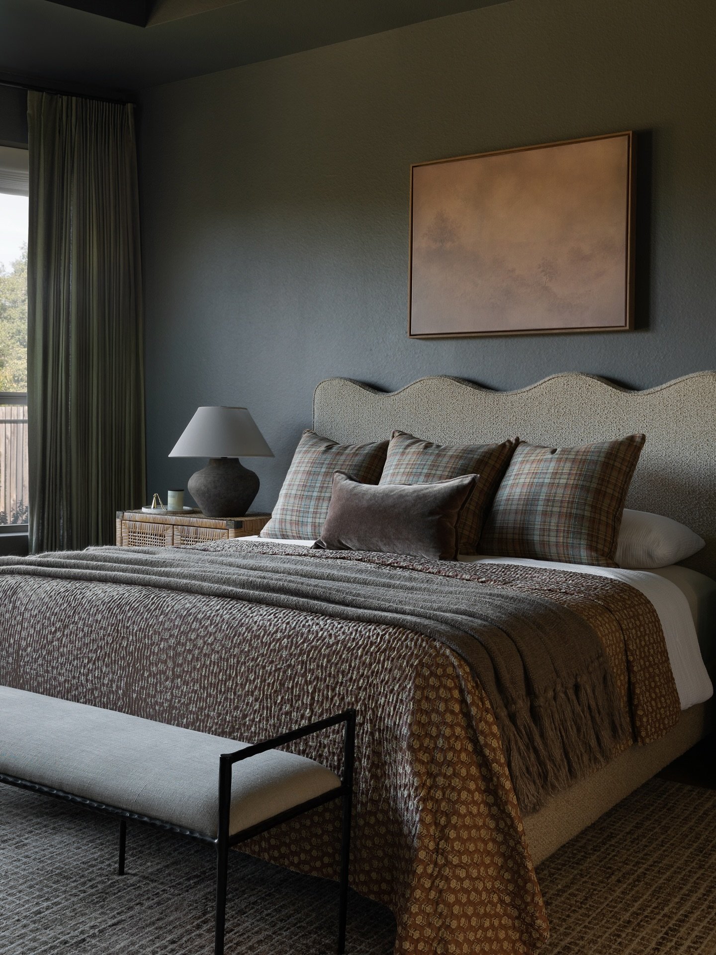 Dark bedroom with matte charcoal grey walls, a scalloped fabric headboard, layered brown and plaid bedding, and a woven nightstand with a ceramic lamp.