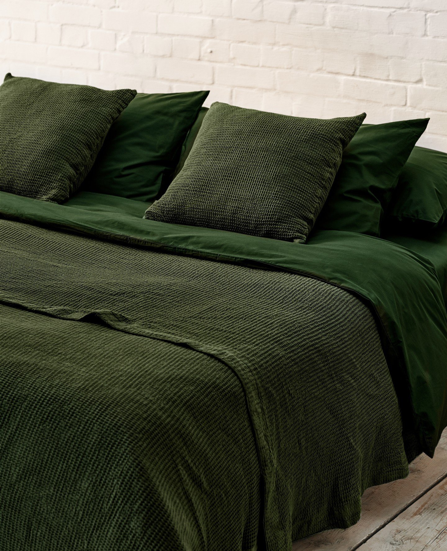 Close-up of a bed layered with rich green velvet sheets, waffle knit pillows, and a textured moss green blanket against a whitewashed brick wall.