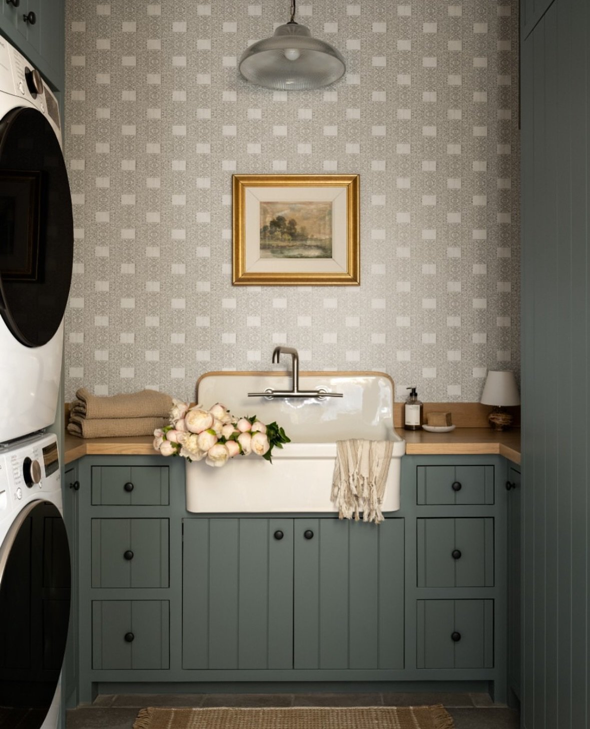 A small, U-shaped laundry room with a stacked washer/dryer on the left and a large, exposed ceramic farmhouse sink centered over deep green-blue cabinets and a light geometric wallpaper.