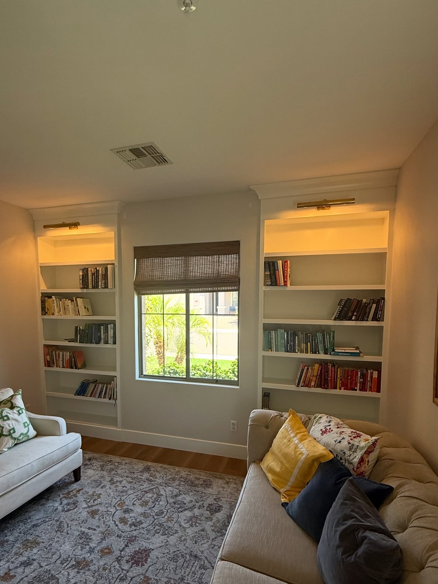 Two identical white built-in bookcases installed symmetrically on either side of a central window, featuring gold picture lights at the top to illuminate the shelves of books.