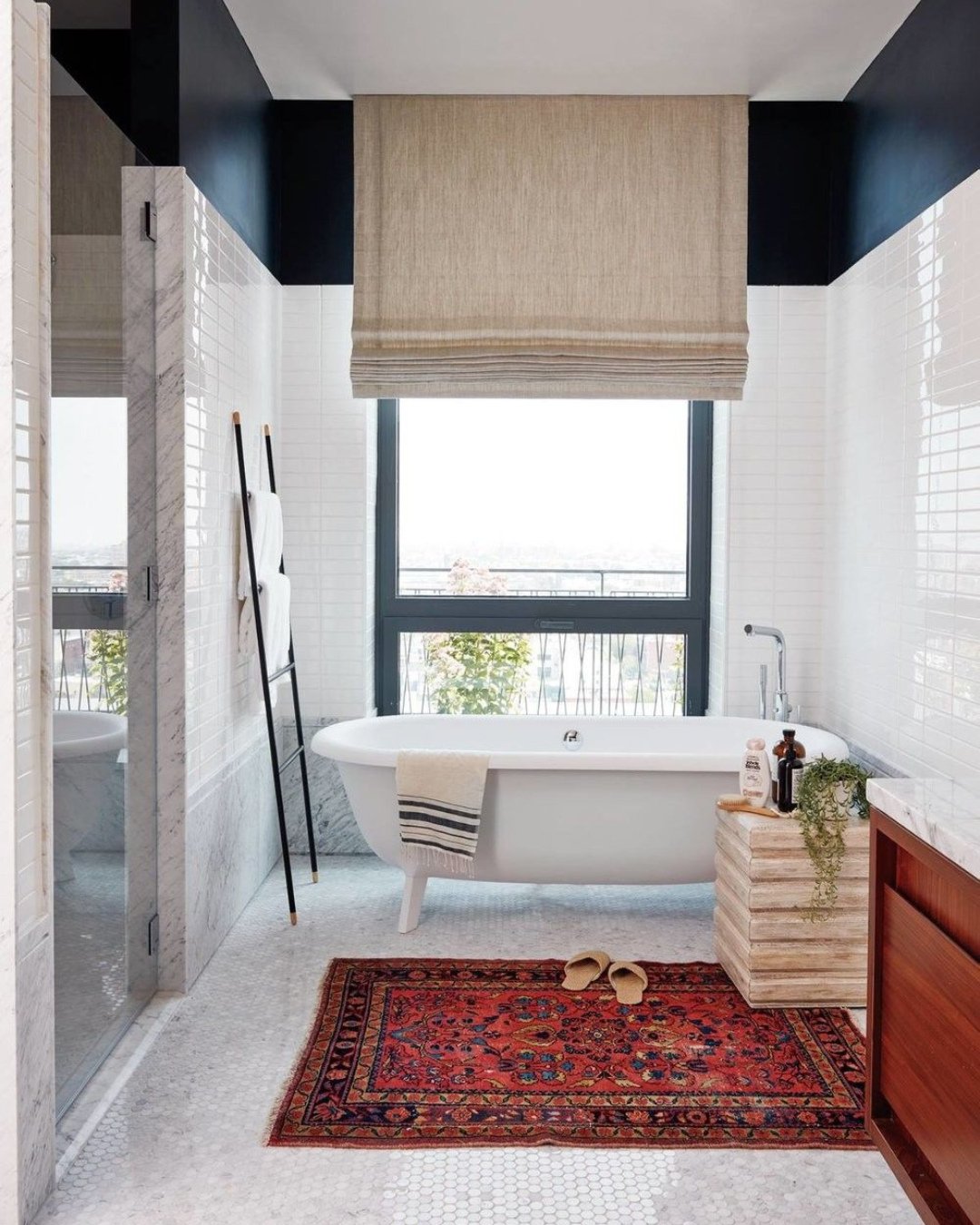 A contemporary bathroom featuring long white subway tile walls, a white hexagonal marble floor, a freestanding tub beneath a window, and a small, vibrant red and blue traditional Persian rug.