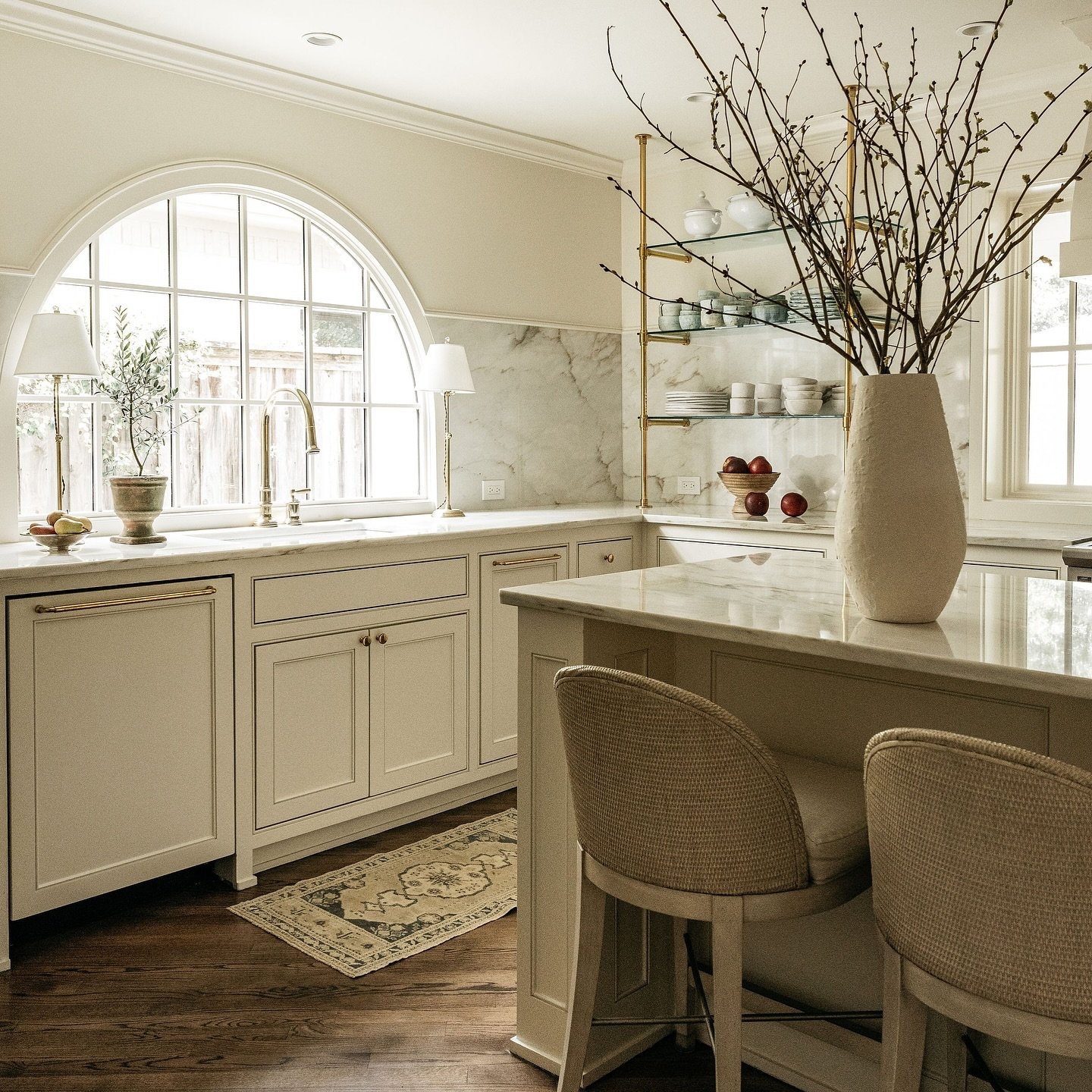Luxurious kitchen with creamy white cabinets and marble counters. A large, dramatic arched grid window is centered over the sink, flanked by matching gold table lamps on the counter.