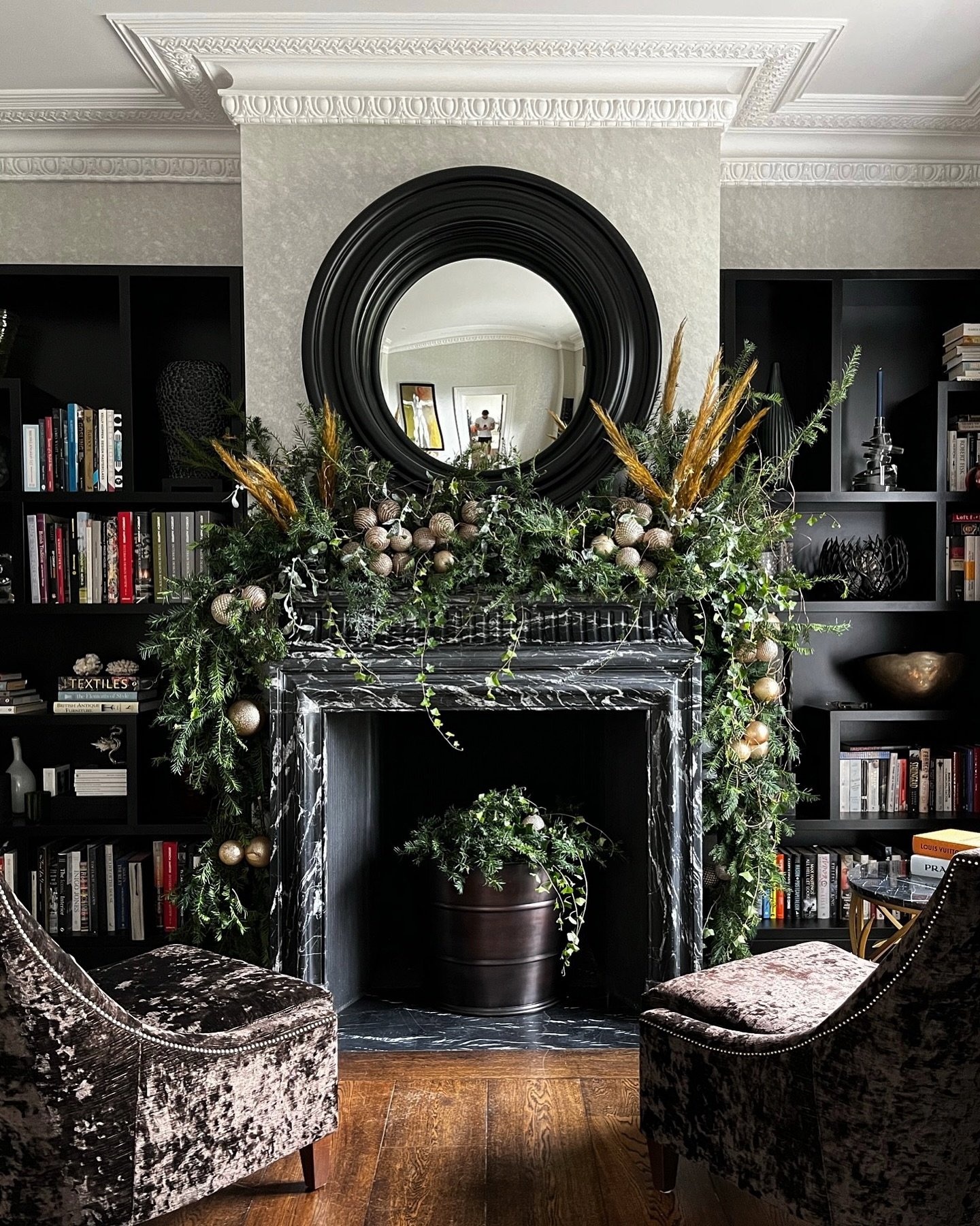 Black marble fireplace and surrounding black bookshelves, heavily decorated with thick, flowing evergreen garland, matte gold ornaments, and tall wheat-colored plumes.