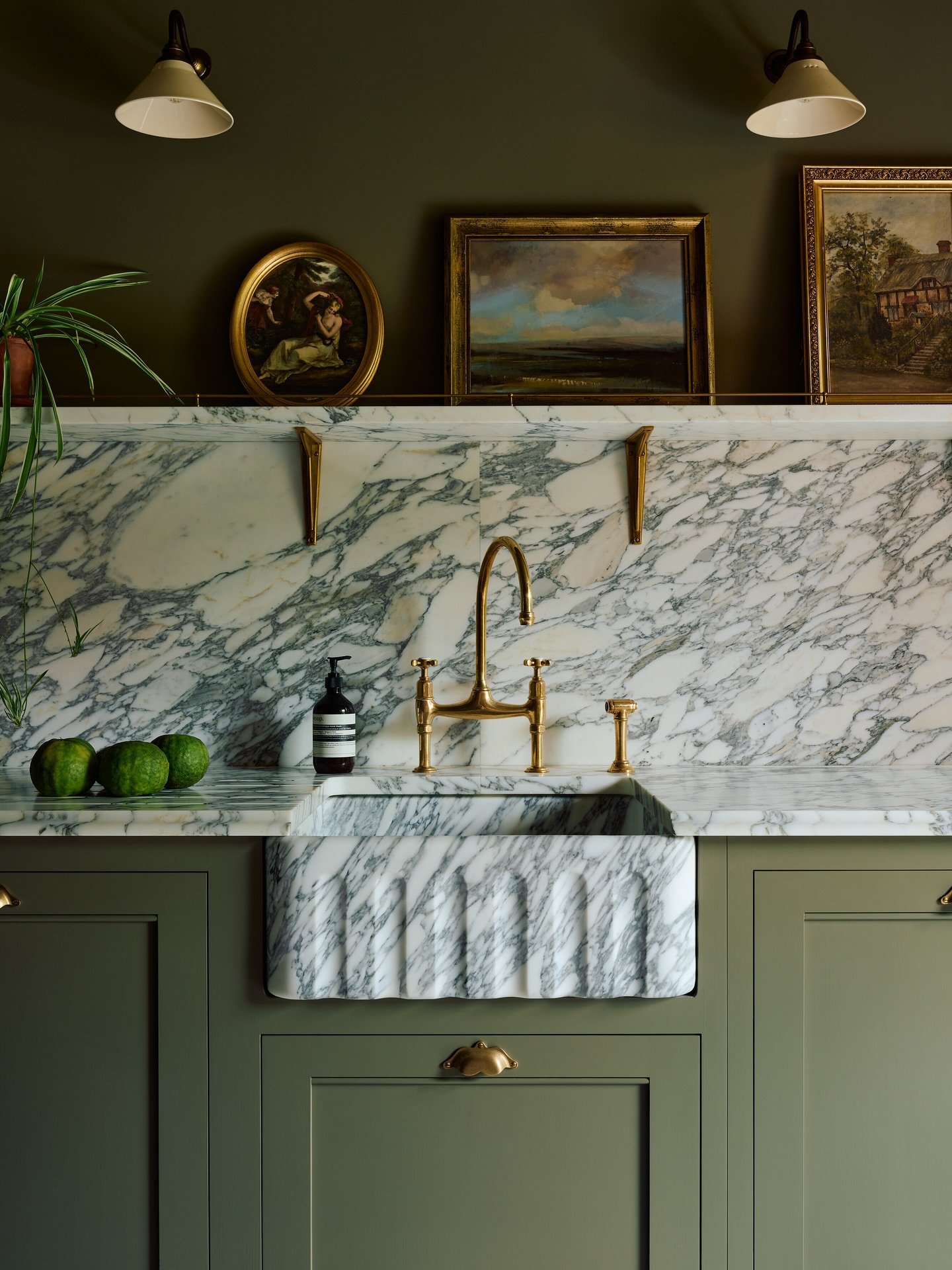 A custom-carved marble farmhouse sink with a decorative scalloped or fluted apron-front, integrated into matching marble countertops and backsplash, set over sage green cabinets with a brass bridge faucet.