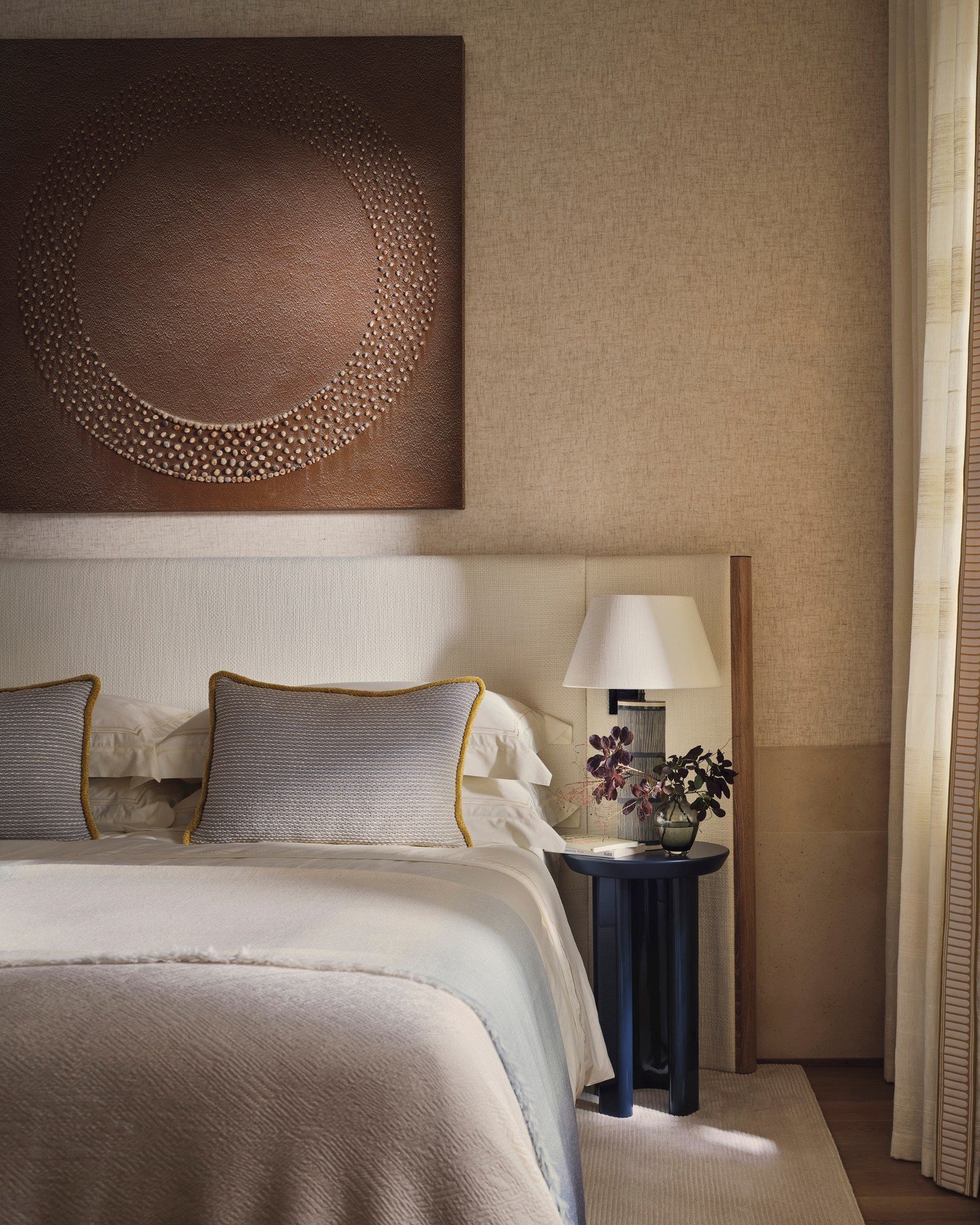 Wide, low upholstered headboard in a light cream textured fabric with a solid wood frame border, sitting beneath large beaded circular wall art.
