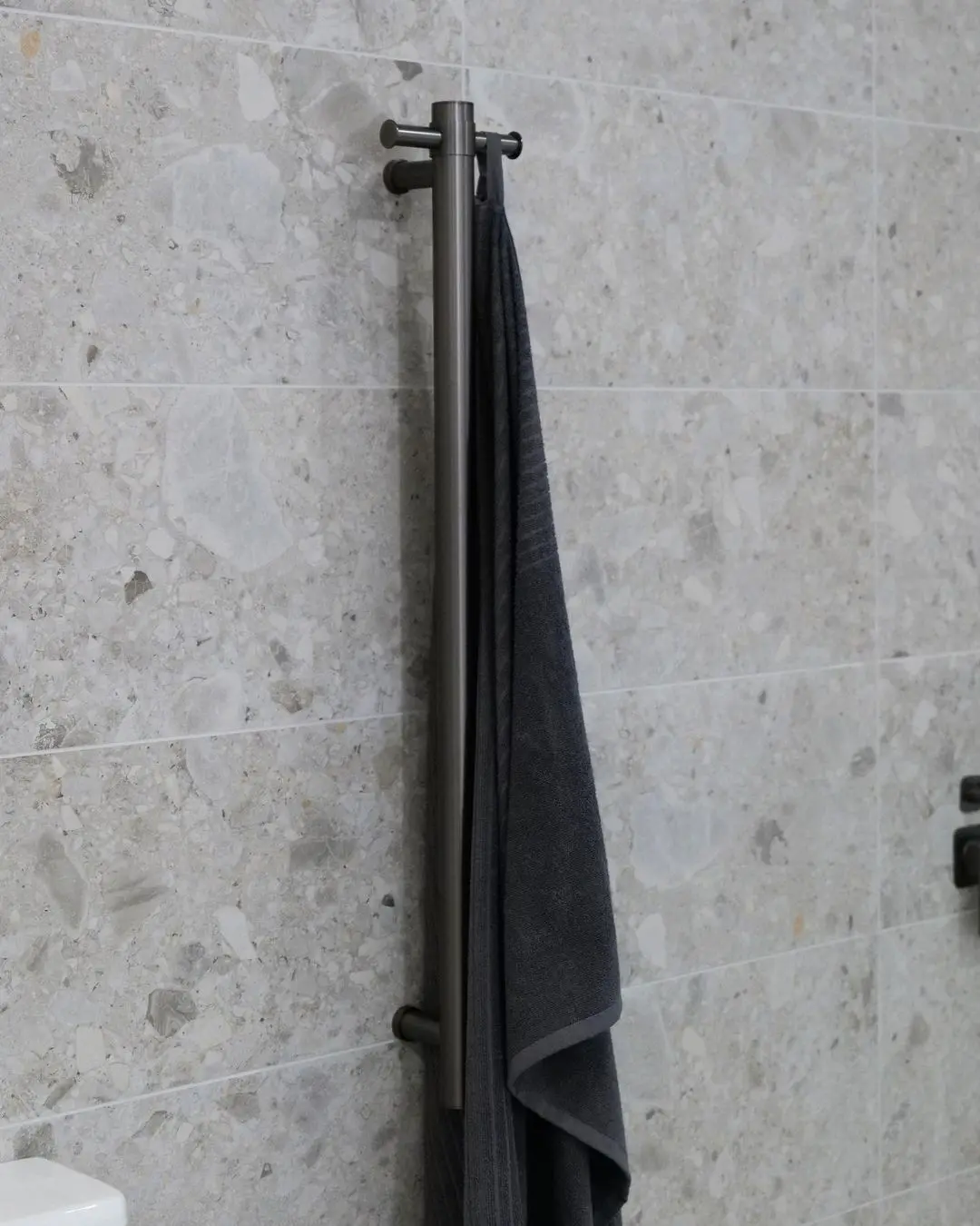 Close-up of a dark gunmetal grey vertical towel rail with small pin-like arms at the top and bottom, holding a dark grey bath towel against a grey terrazzo-look tile wall.