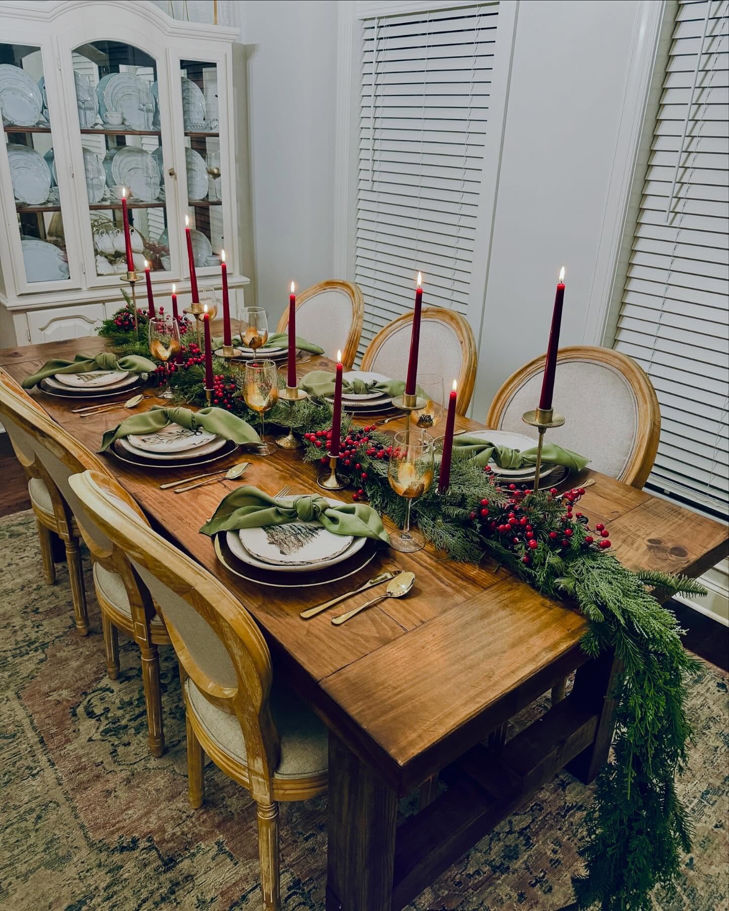 A rustic wood dining table with elegant linen-backed chairs, featuring a long centerpiece of evergreen garland, red berries, and seven tall burgundy taper candles in brass holders. Place settings have gold chargers, olive green napkins, and small decorative plates.