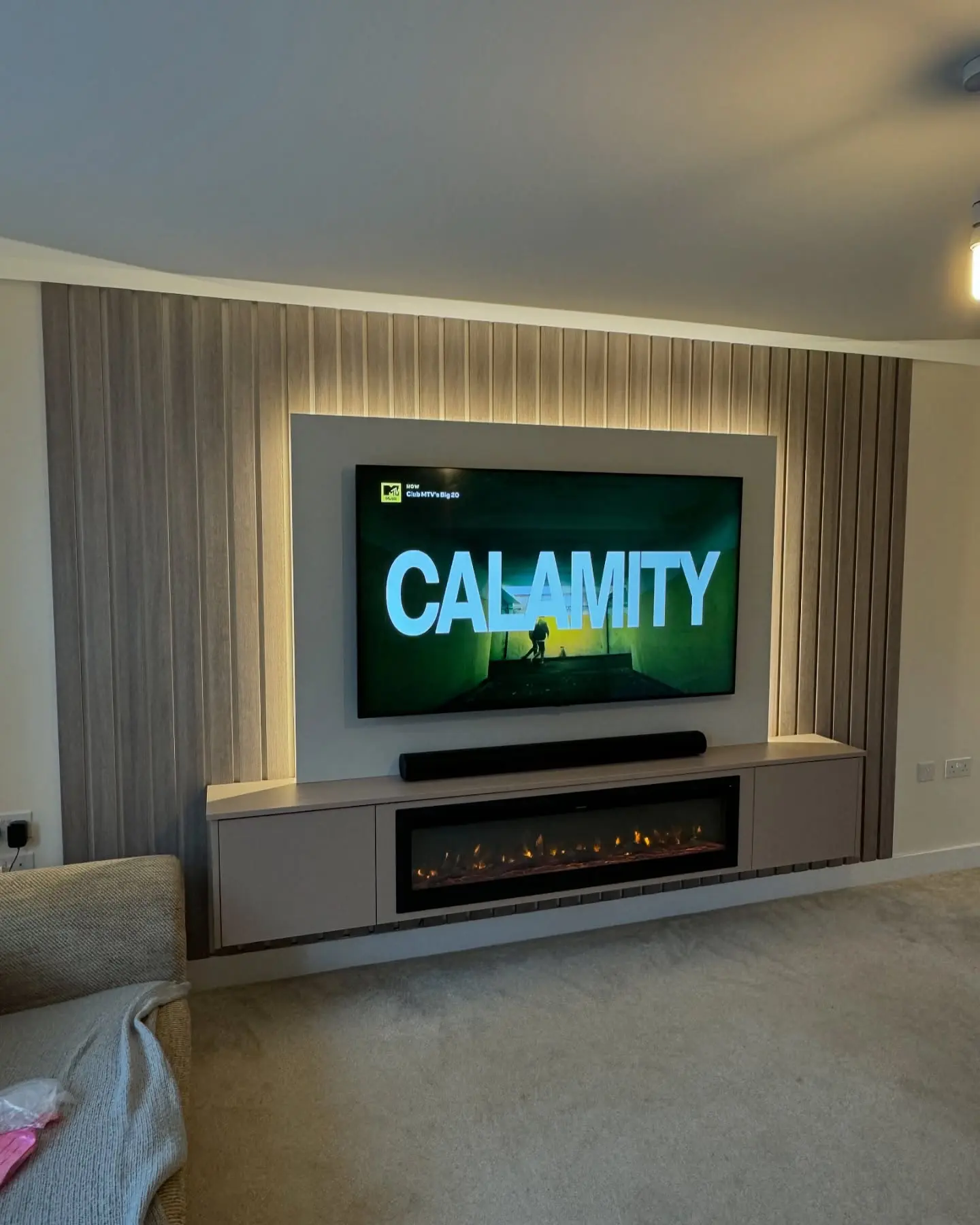 Modern media wall with vertical slat paneling, a backlit recessed panel for the TV, and a floating taupe console housing a long linear electric fireplace.