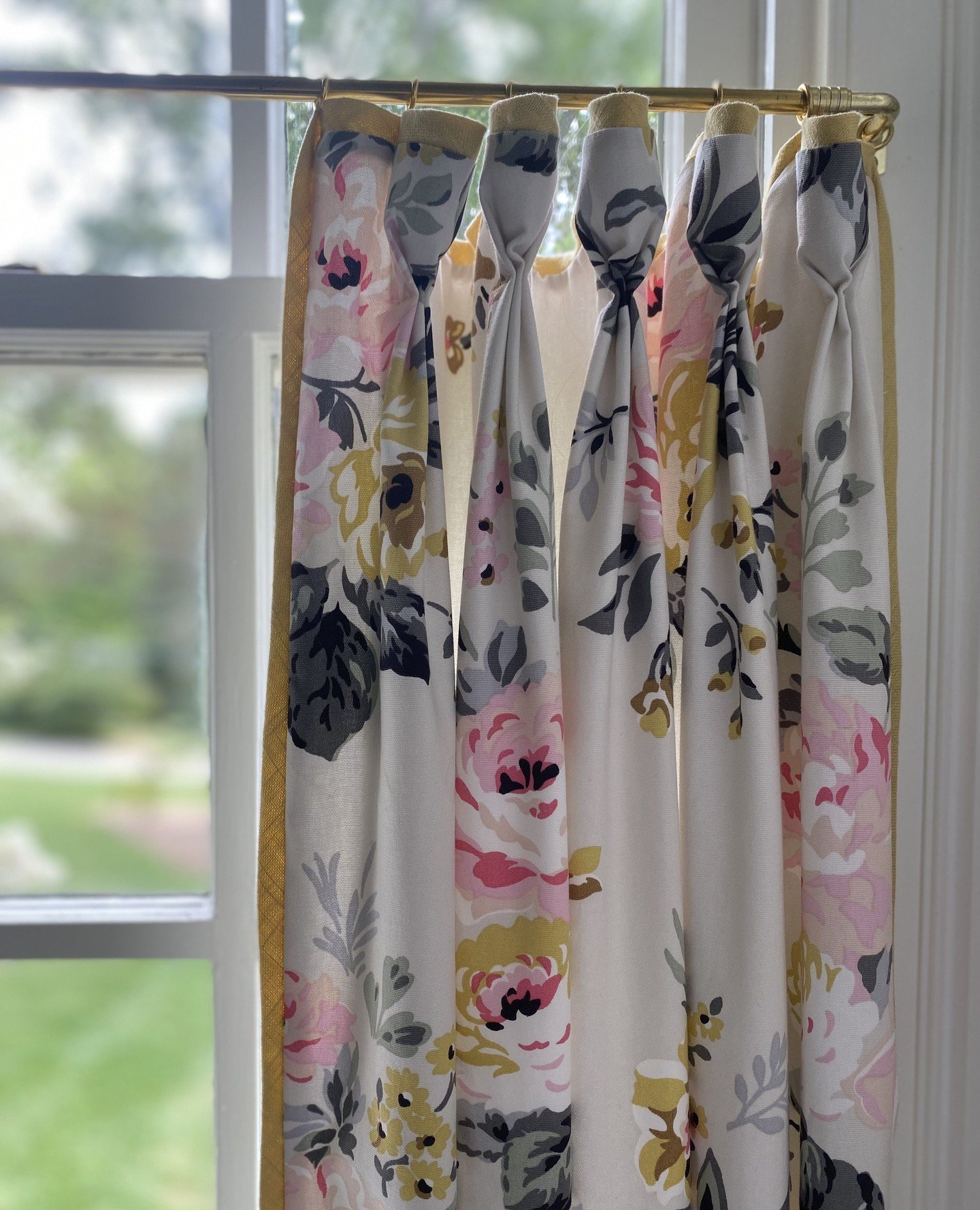 Close-up of a kitchen curtain panel with a pink, yellow, and gray floral print, hung on a brass rod using decorative fabric tie tops.