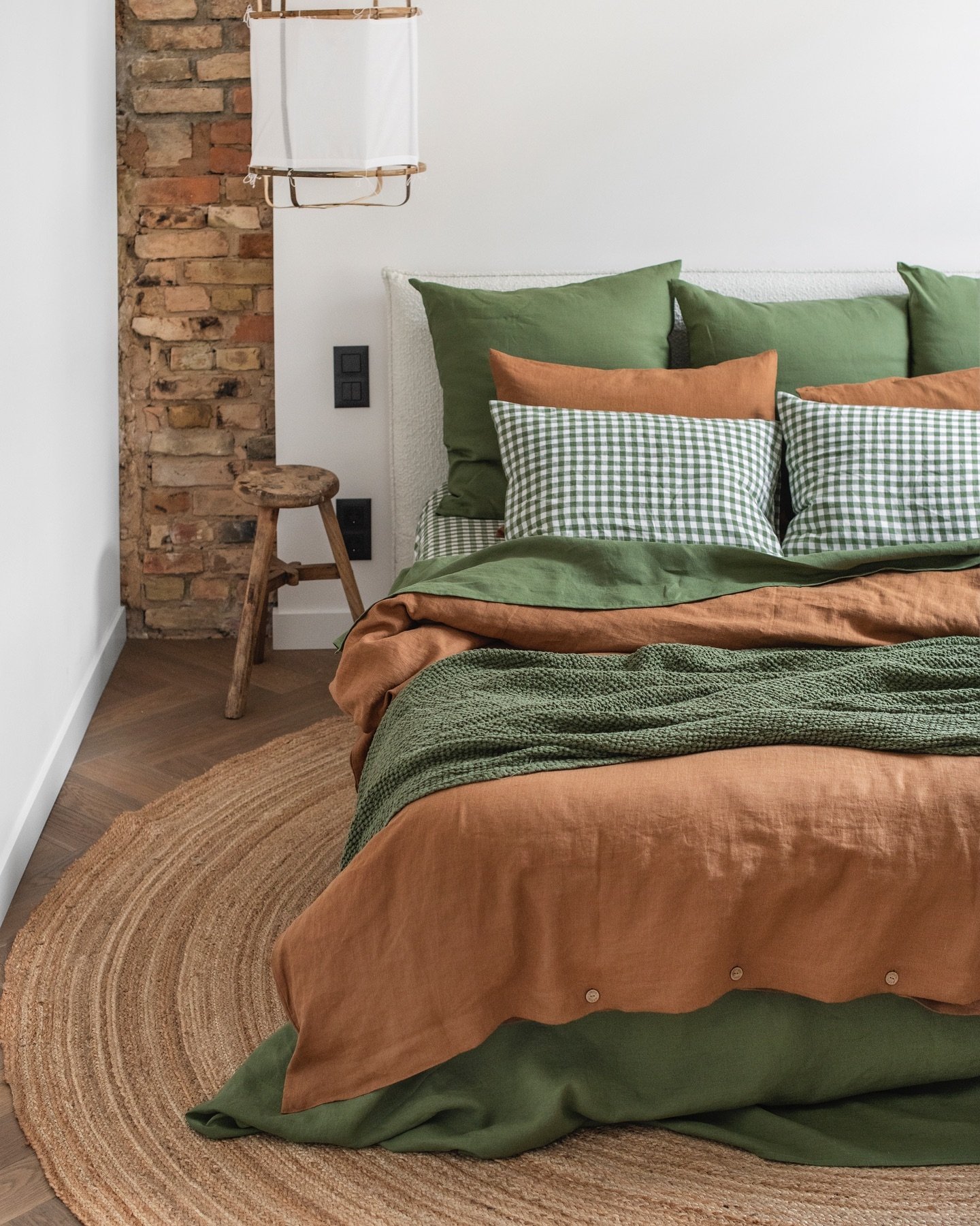 Close-up of a bed with layered deep green and terracotta linen bedding, featuring a jute rug and exposed brick.