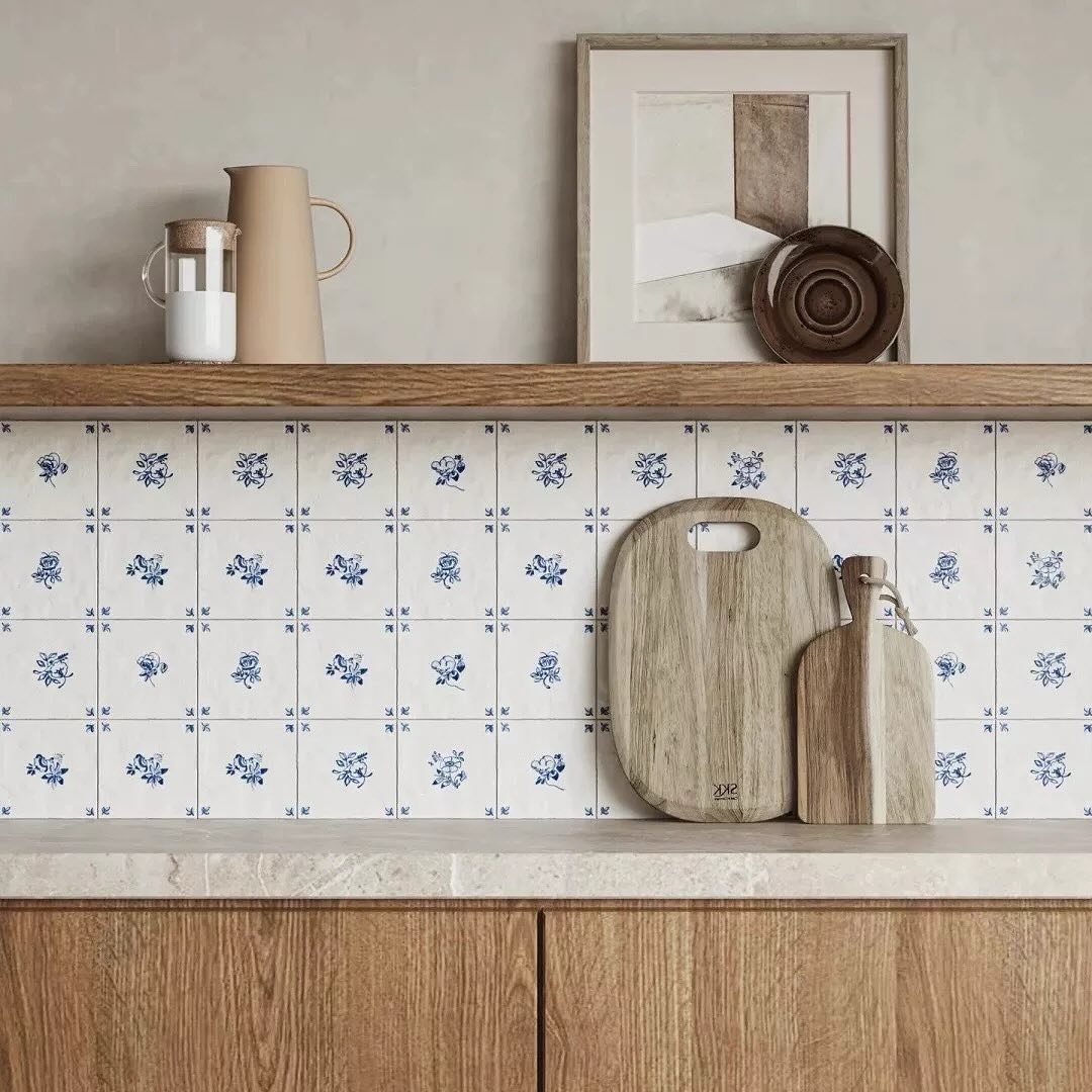 Kitchen backsplash of small square white tiles with a repeating hand-painted blue floral motif, set beneath a wooden shelf and above natural wood cabinetry.