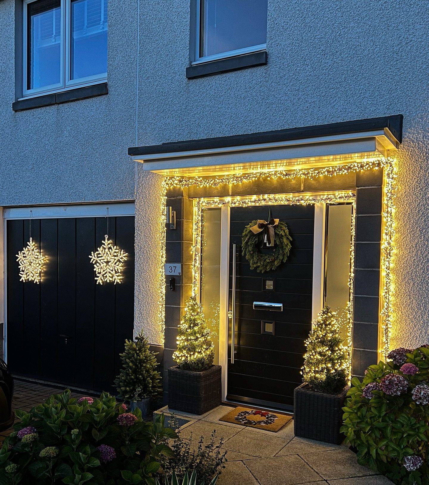 A contemporary home entryway is framed by thick strands of warm white icicle lights around the door, with two small lighted trees flanking the black door, and two snowflake motifs hanging on the black garage doors.