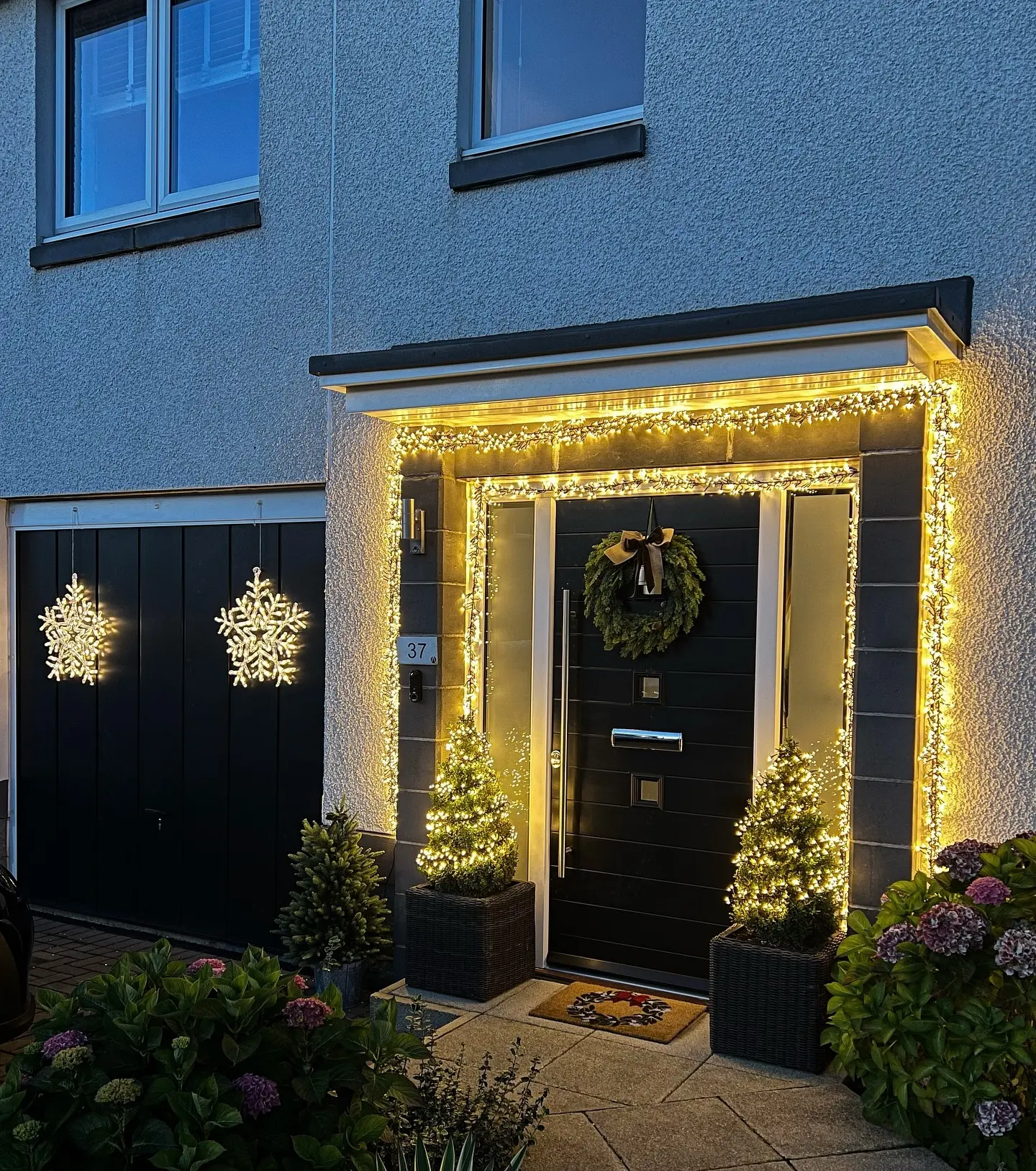 A contemporary home entryway is framed by thick strands of warm white icicle lights around the door, with two small lighted trees flanking the black door, and two snowflake motifs hanging on the black garage doors.