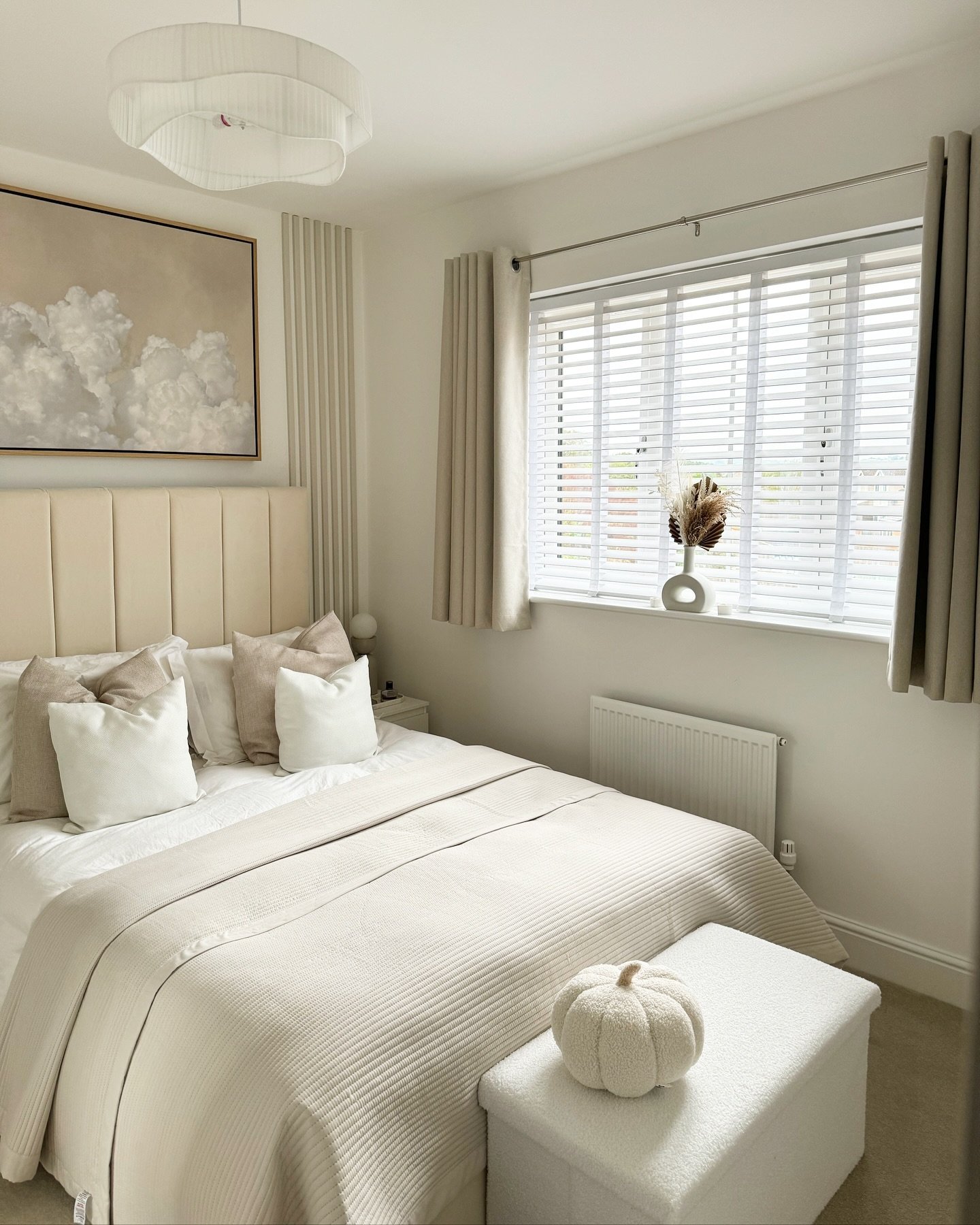 A bright beige bedroom with a fluted headboard, ribbed throw blanket, a white upholstered storage ottoman, and abstract artwork depicting soft clouds.