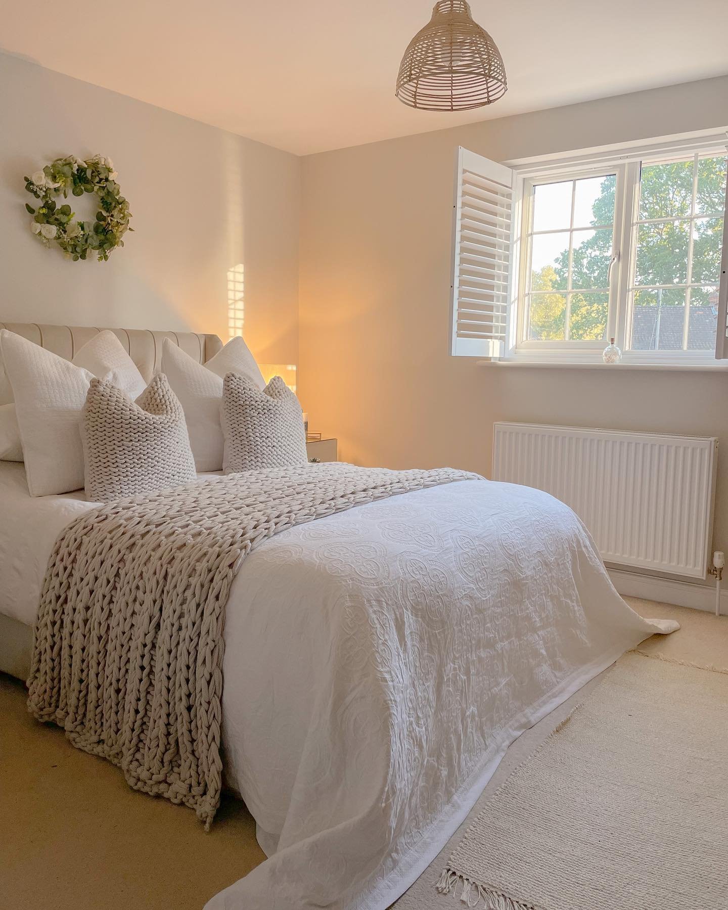 Bright bedroom with white window shutters, a white duvet with an embroidered pattern, a chunky knit beige throw, and matching knitted cushions.