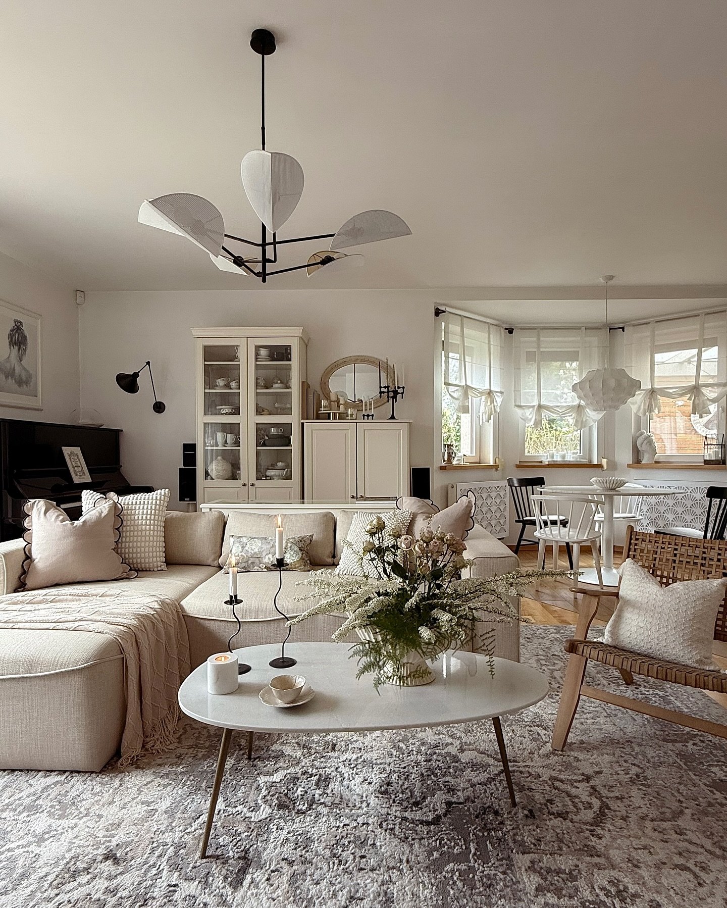 A cozy living room featuring a light beige sectional sofa, a vintage china cabinet, a white oval coffee table, and a sculptural modern chandelier.