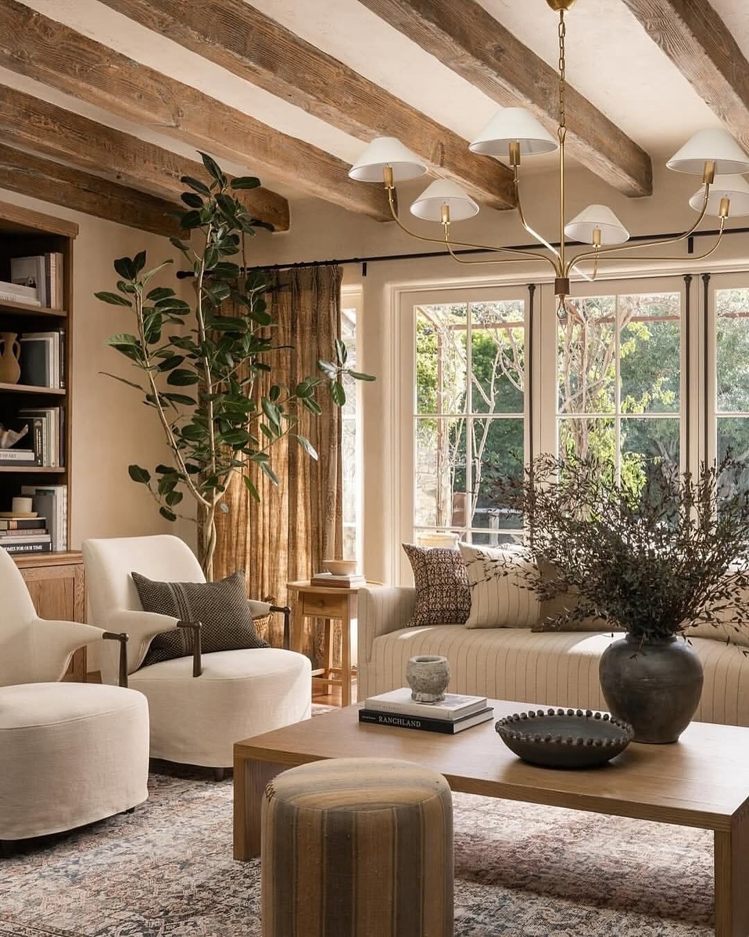 A cozy beige living room with dark wood ceiling beams, cream-colored barrel chairs, a striped sofa, and a large window overlooking a garden.