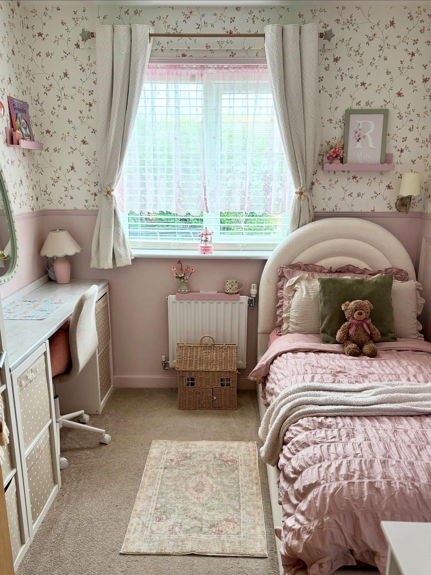 Small child's bedroom featuring floral wallpaper, pink half-wall paneling, a small writing desk, and a rattan basket.