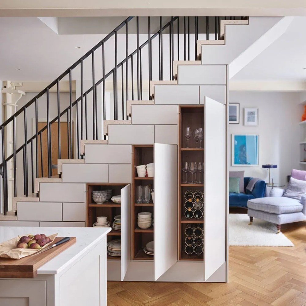 White, tiled-look storage cabinets under open-tread stairs, featuring wooden shelves for crockery.