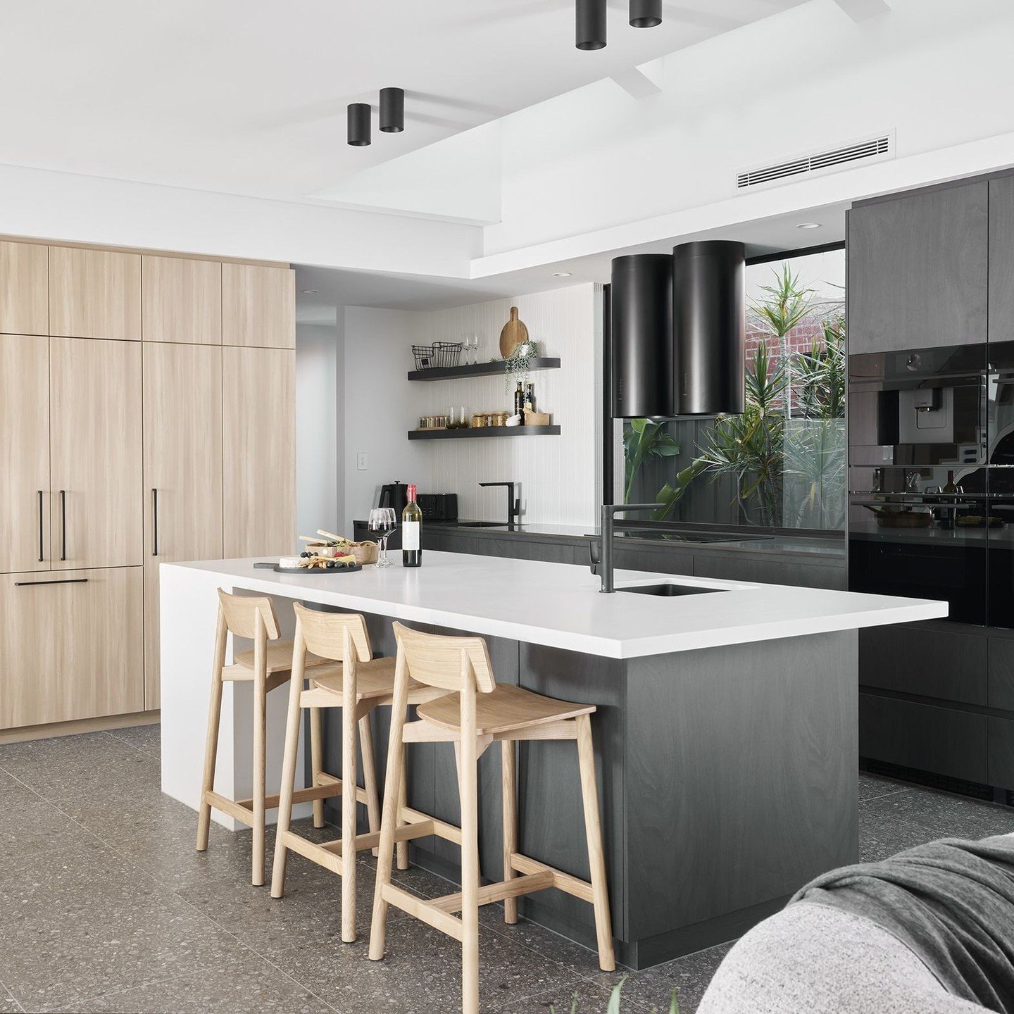 Open-plan modern kitchen with light wood full-height cabinets, a dark grey island with a white countertop, and distinctive black cylindrical range hoods.
