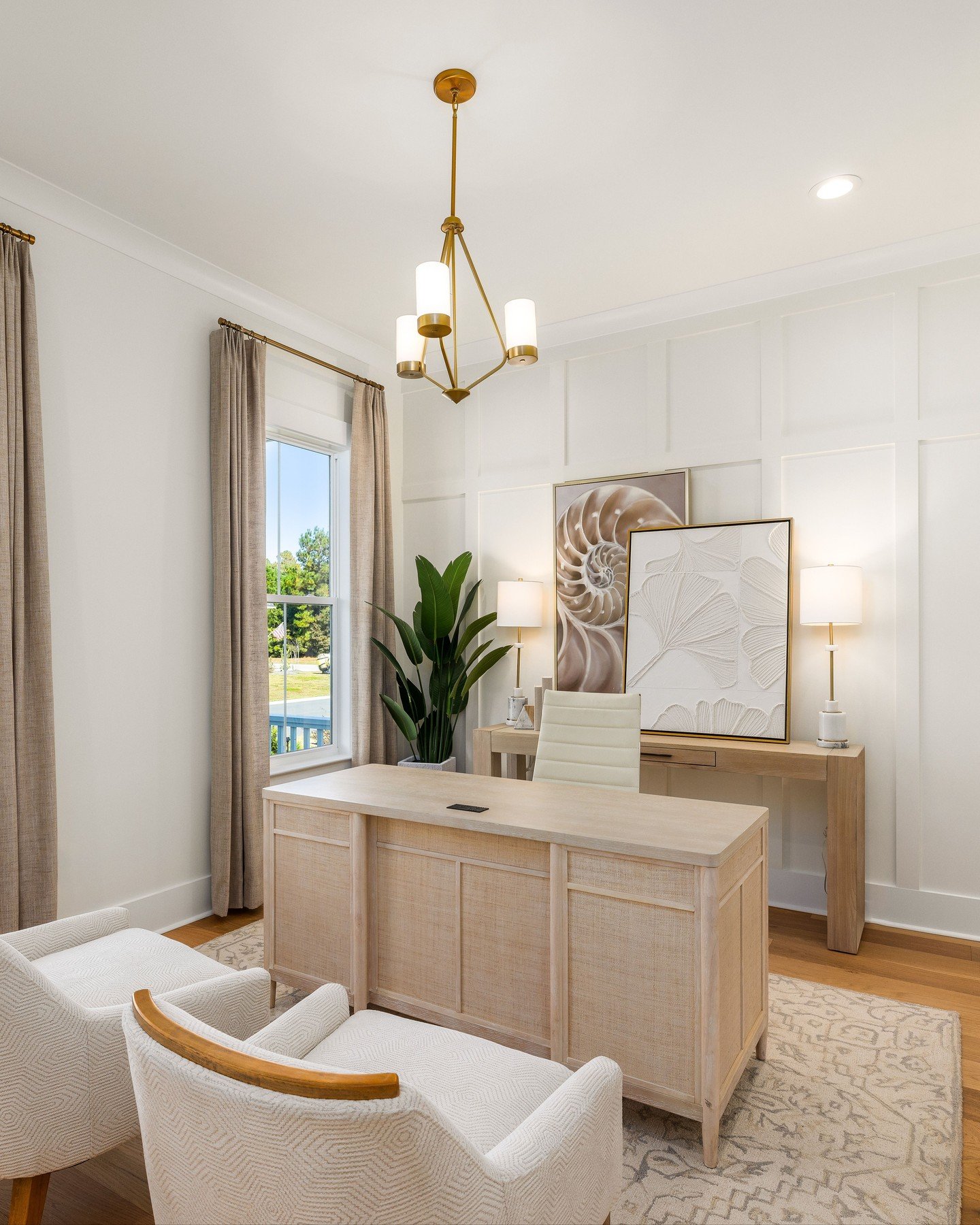 Bright, coastal-style home office with a light wood and cane executive desk, white paneling on the wall, and a gold geometric chandelier.