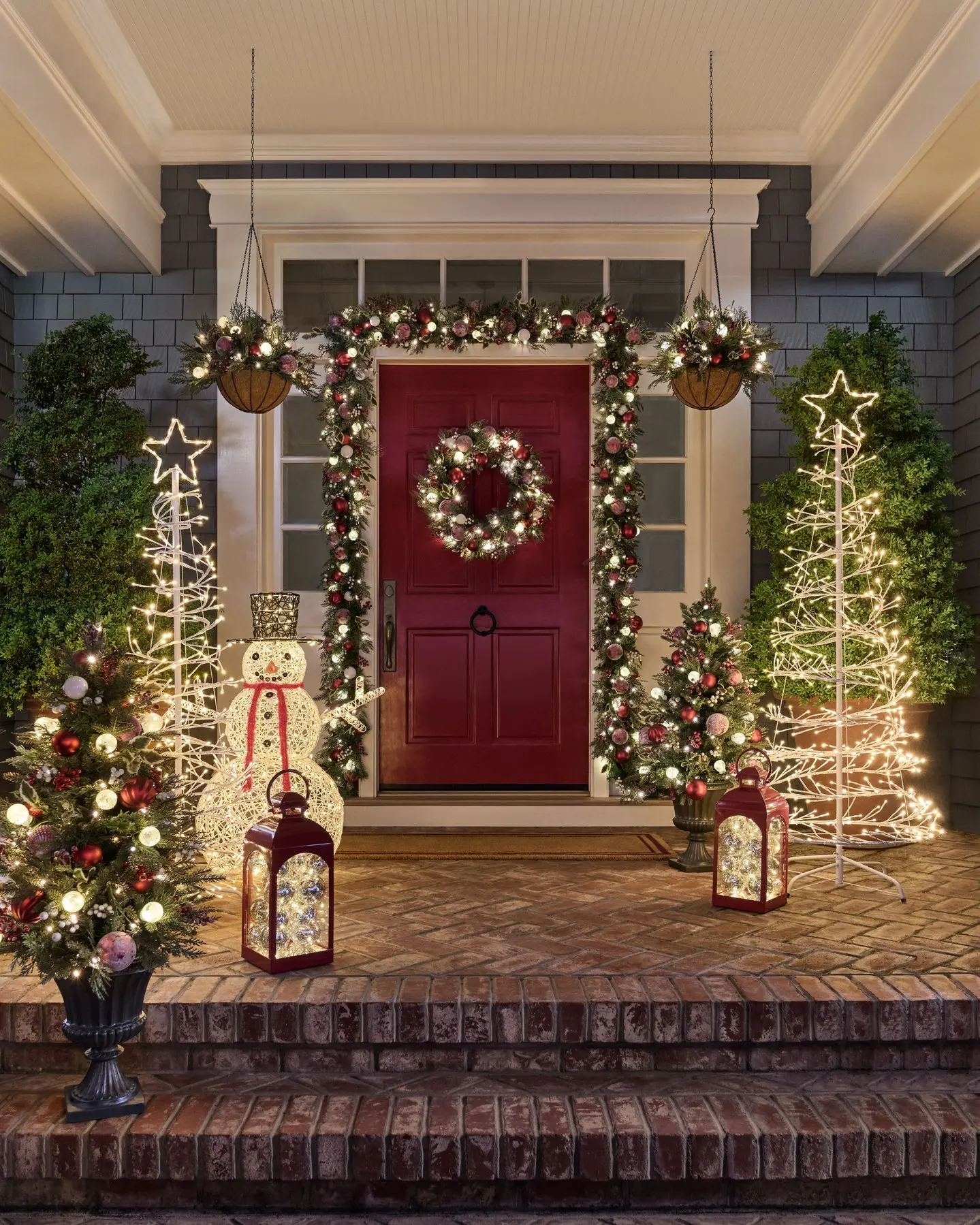 A vivid red front door is surrounded by a thick garland with red accents, a lighted wreath, a glowing wire-frame snowman, lighted tree motifs, and two red decorative lanterns.