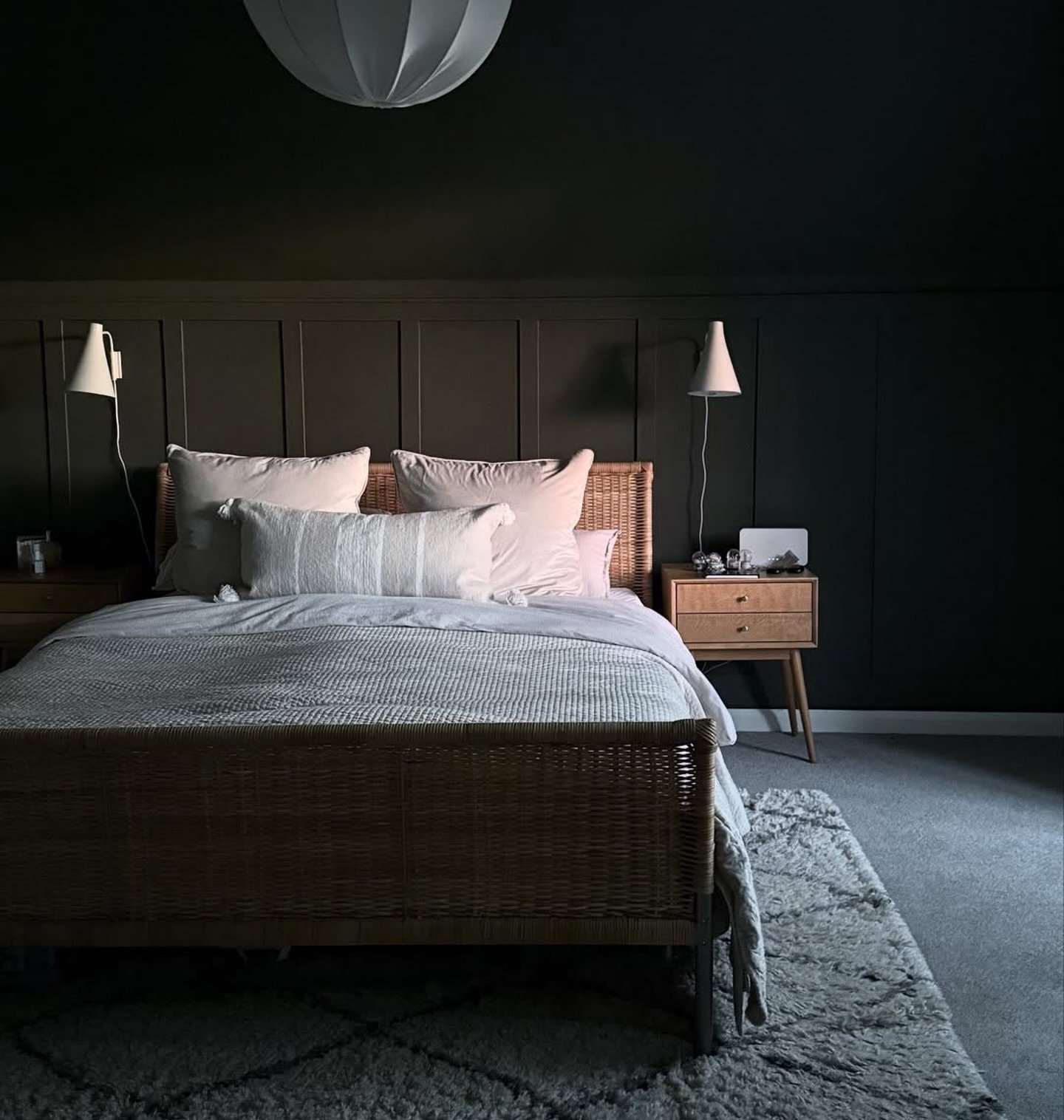 Dark bedroom with matte black walls, a black paneled accent wall, a light wicker bed frame, white cottagecore bedding, and mid-century nightstands.