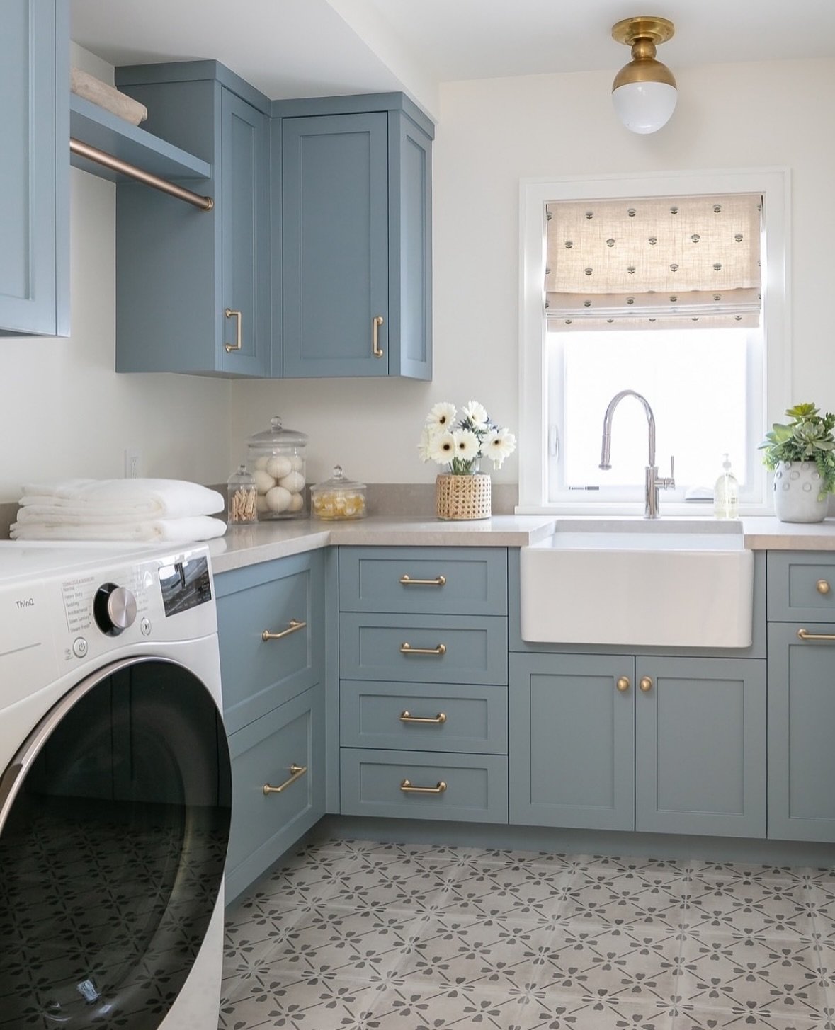 A small, bright laundry room featuring extensive dusty blue cabinetry, a white farmhouse sink centered under a window, brass hardware, and a floor covered in a gray floral cement tile.