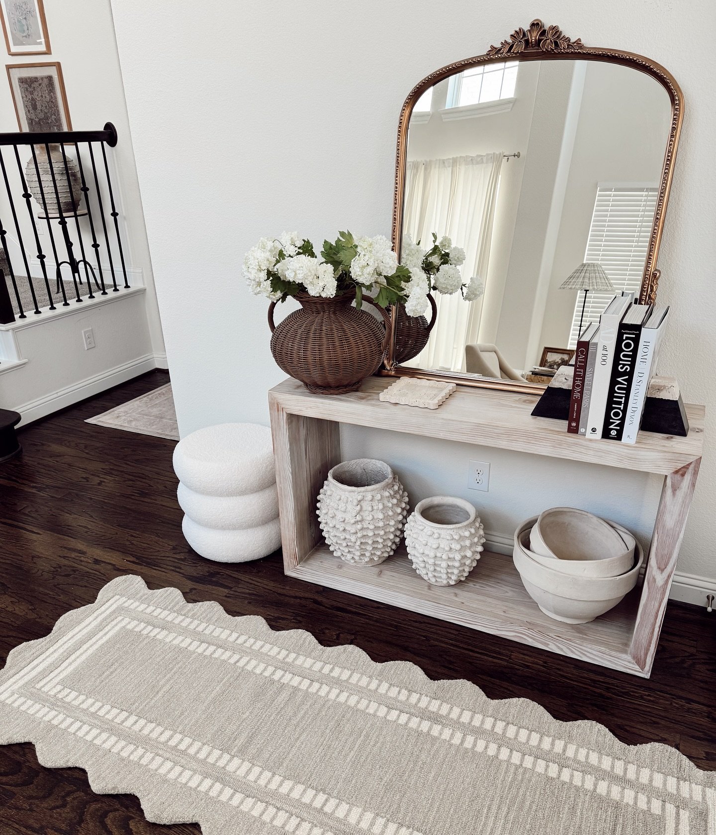 A bleached wood open console shelf with a large ornate gold mirror, a woven vase, and decorative white pottery and books on a dark wood floor.