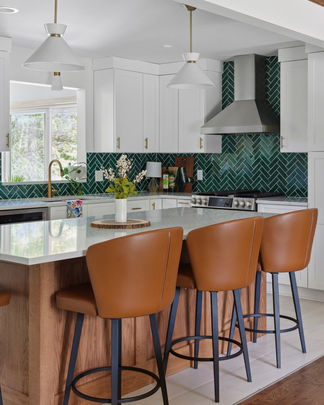 Modern white kitchen with a vibrant glossy teal or dark green subway tile backsplash installed in a herringbone pattern, paired with wood accents and tan leather bar stools.
