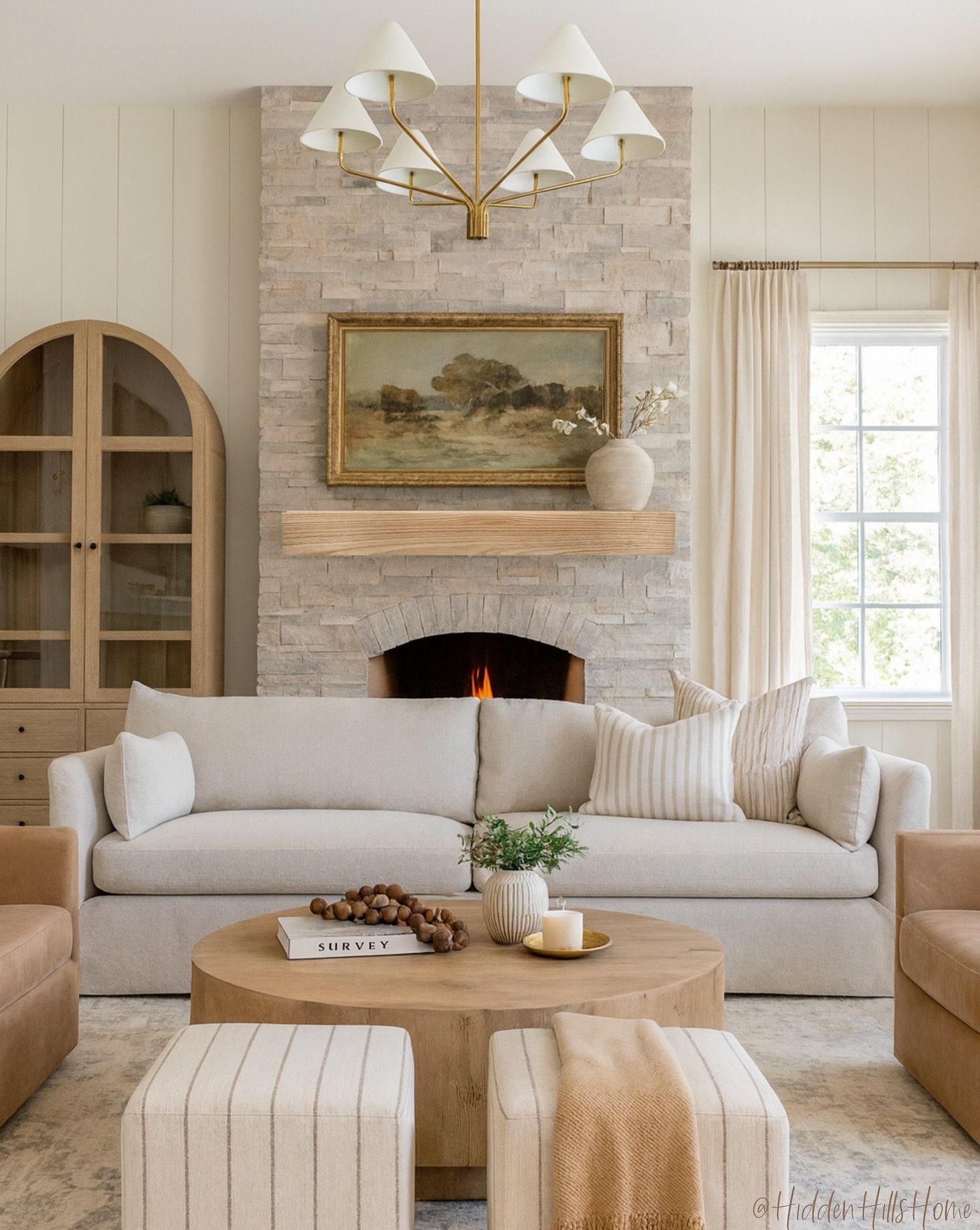 A cozy, light-filled living room featuring a large, stacked natural stone fireplace, a gold mid-century modern chandelier, a light linen sofa, and an arched wood cabinet.