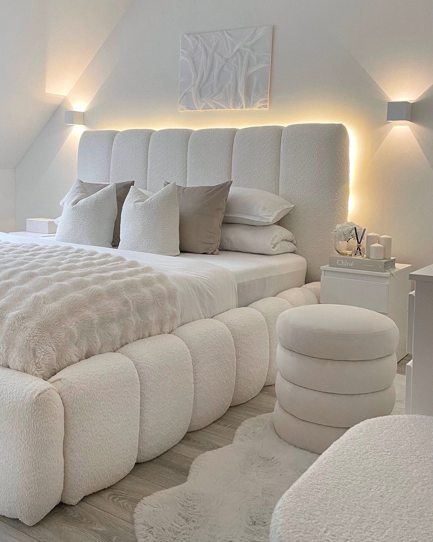 All-white bedroom featuring a large channel-tufted bouclé bed frame with under-bed lighting, a circular stacked ottoman, and two white wall sconces.