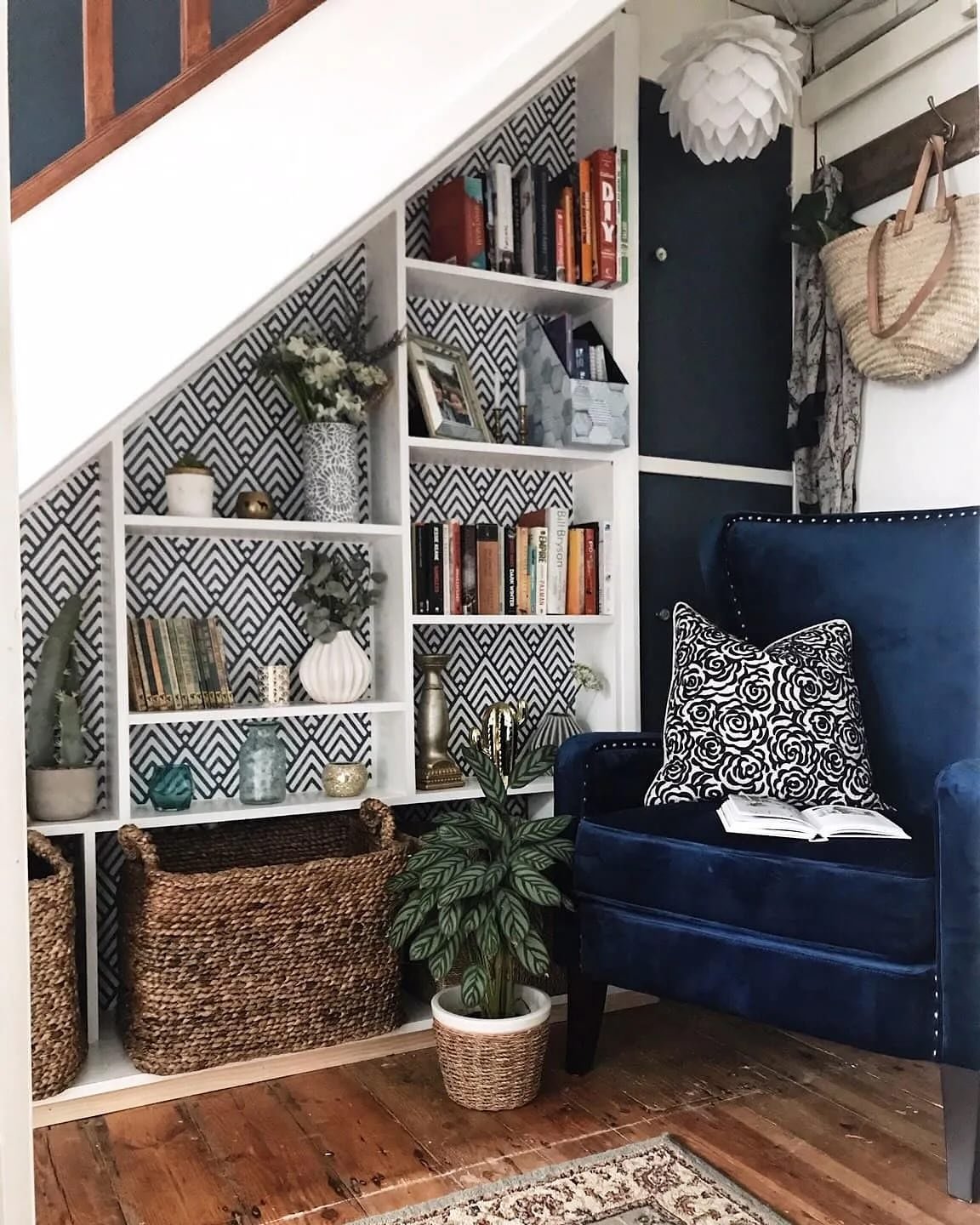 White open shelves with patterned wallpaper under stairs next to a blue velvet armchair.