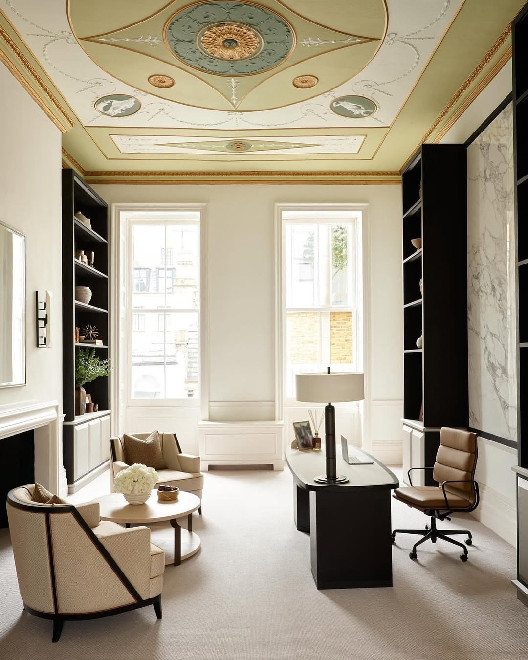 Luxurious classic home office with an ornate painted ceiling, black built-in bookshelves, a dark executive desk, and two light fabric armchairs.