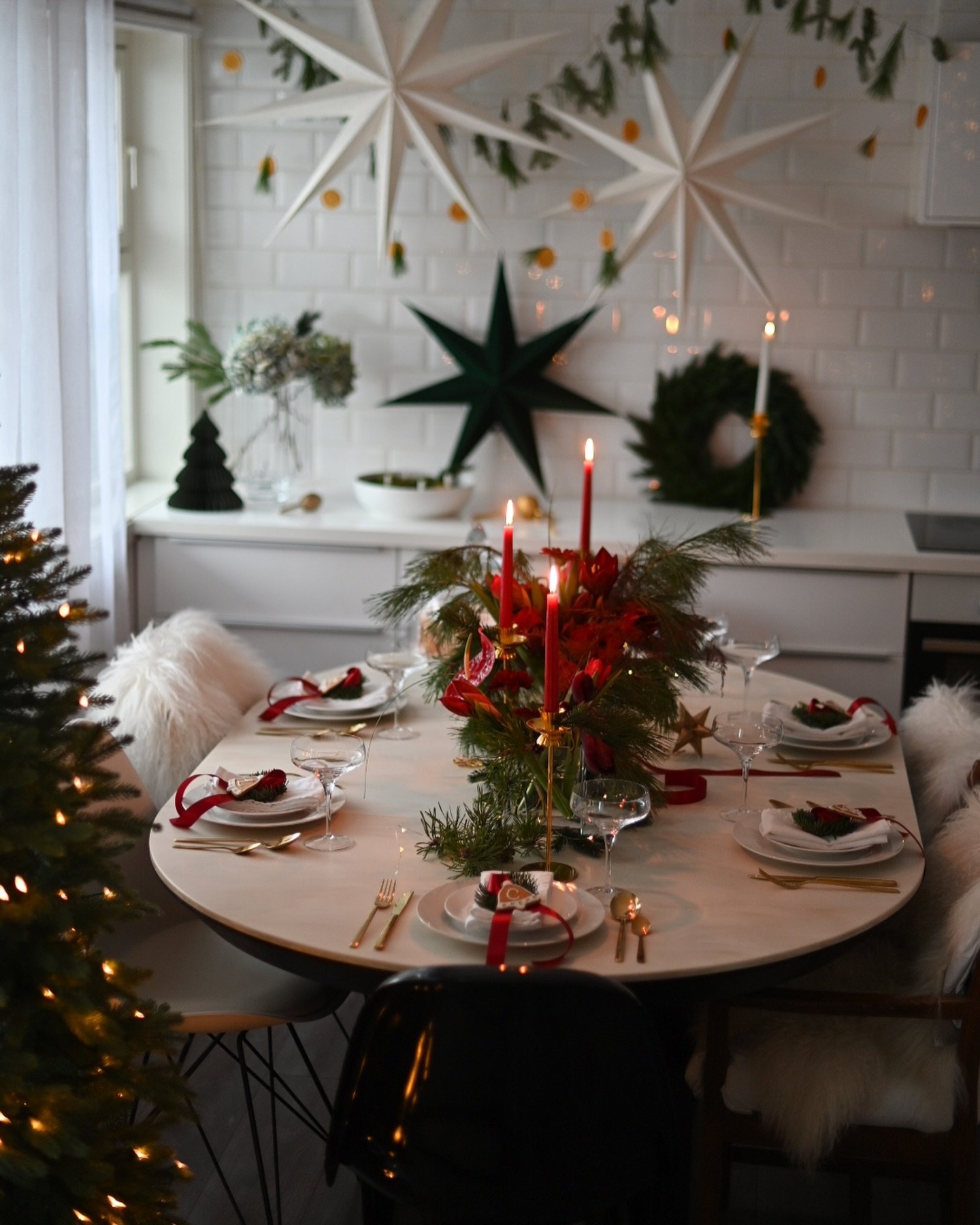 A small round dining table with white sheepskin chair covers, set with layered white and dark plates, gold flatware, and a striking centerpiece of deep red flowers, pine boughs, and crimson taper candles in gold holders. Large white paper stars hang above.