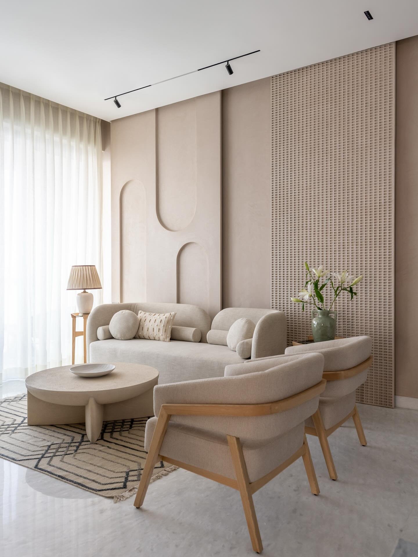 A modern beige living room featuring an artistic sculpted plaster wall, a round sofa, neutral wood-framed armchairs, and a beige stone coffee table.