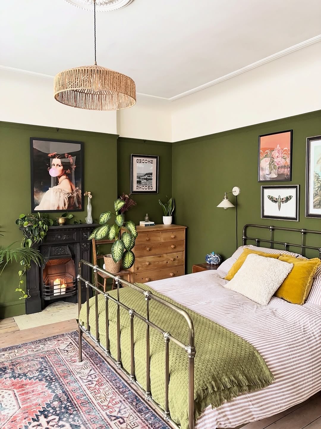 Eclectic bedroom with olive green walls, a brass metal bed frame, a lit fireplace, and a mix of colorful artwork.