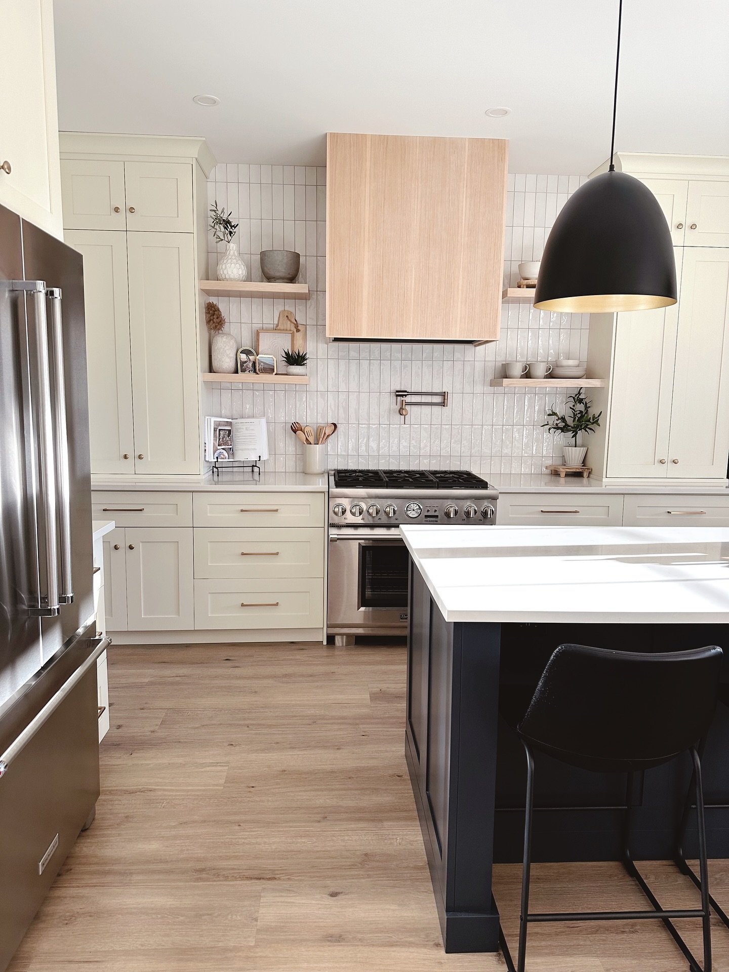 Modern farmhouse kitchen with vertically stacked white subway tile backsplash, a light wood range hood, open floating shelves, and creamy white perimeter cabinets with a dark navy island.