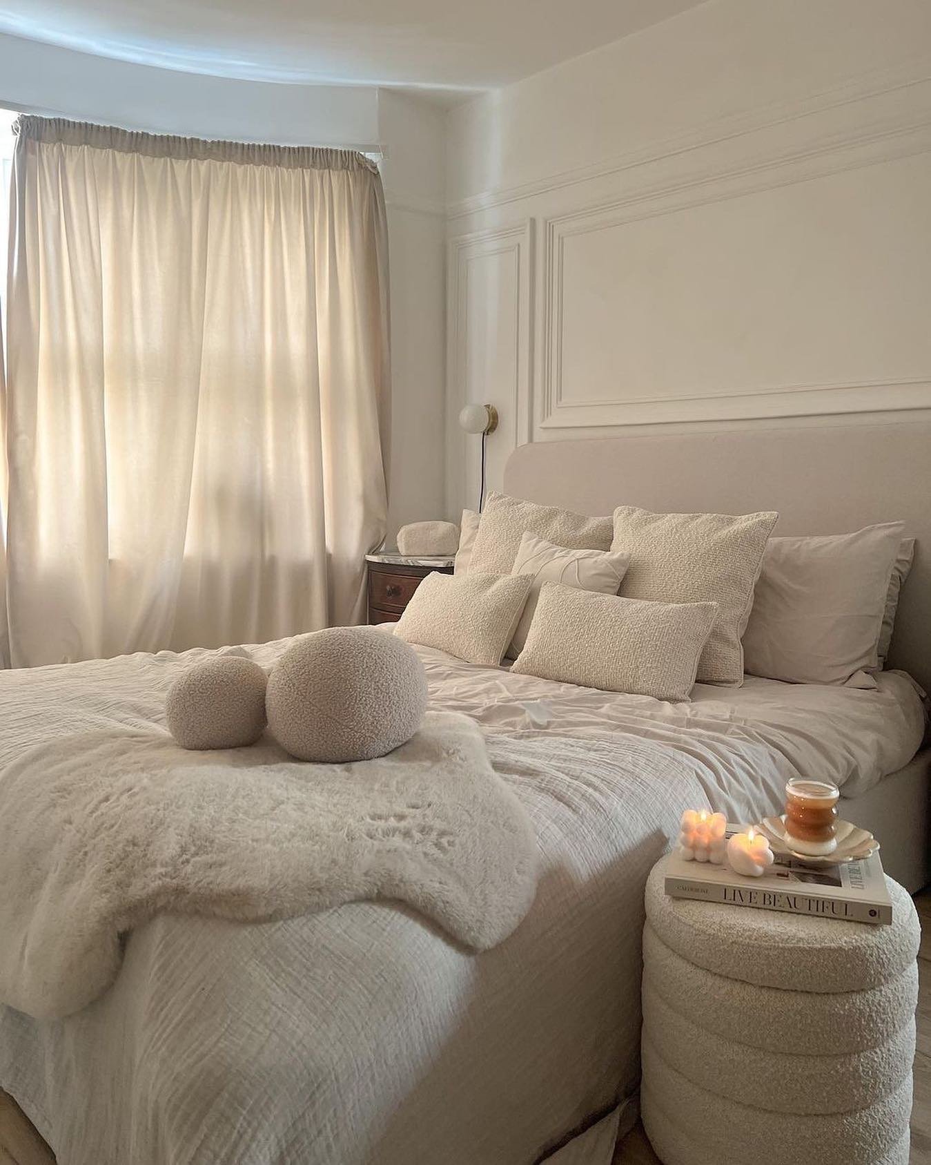 A cozy, neutral beige and white bedroom with a layered bed, a textured sheepskin throw, and multiple round and rectangular cushions in fuzzy bouclé fabric, set against classic wall molding.