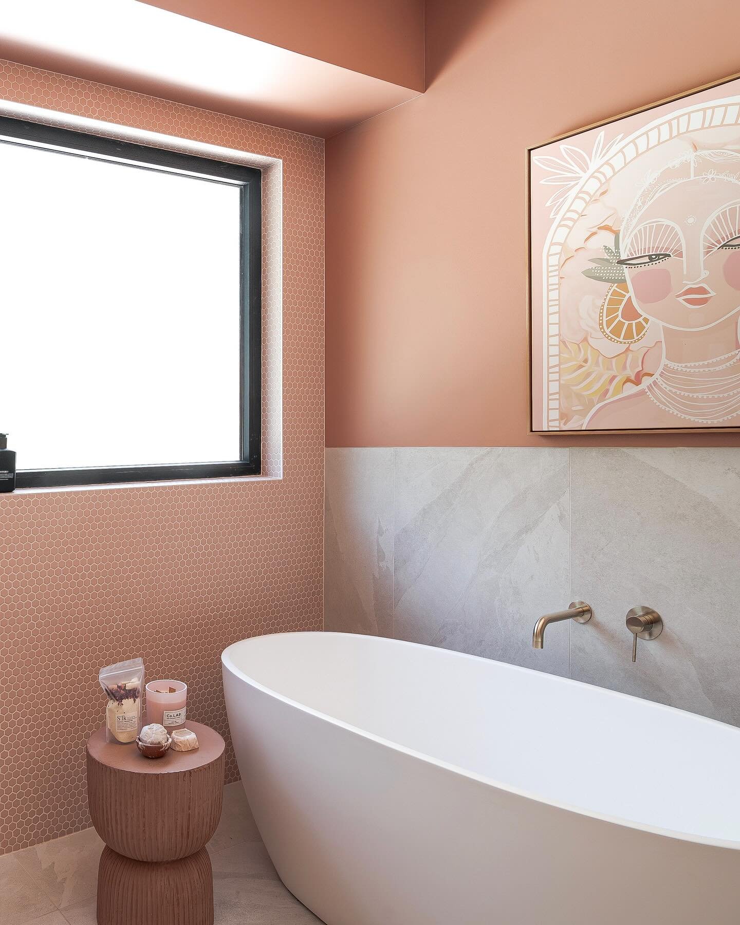 A contemporary bathroom with terracotta-pink walls and gray marble tiles, featuring a large, pale pink and gold portrait painting above a white soaking tub.