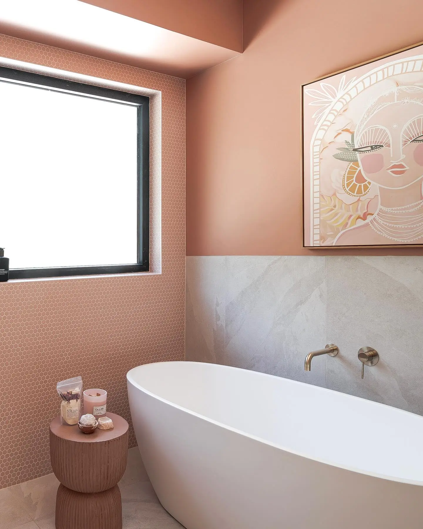 A contemporary bathroom with terracotta-pink walls and gray marble tiles, featuring a large, pale pink and gold portrait painting above a white soaking tub.
