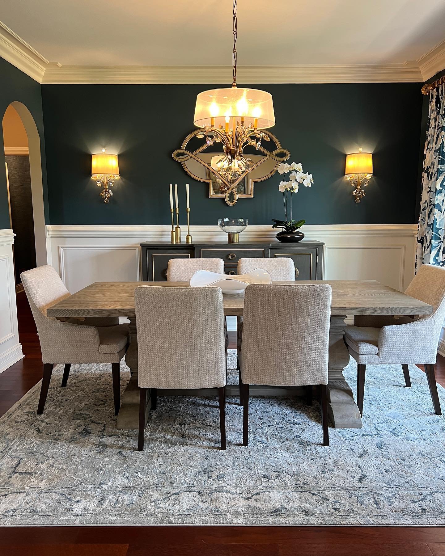 A formal dining room with a dark teal accent wall above white wainscoting, featuring a light wood table, neutral upholstered chairs, and gilded brass sconces and mirror.