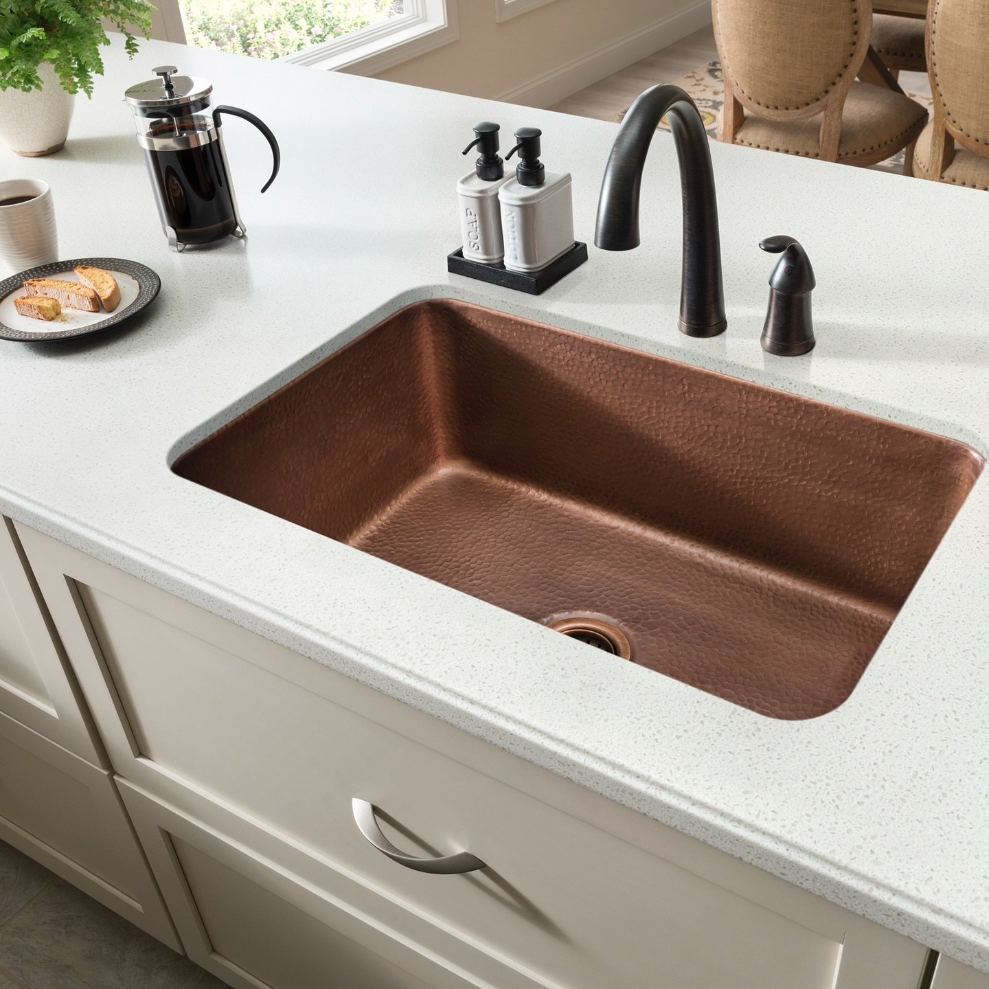 A rectangular single-bowl undermount sink with a hammered, reddish-brown copper finish, set into a white speckled quartz countertop, featuring a dark oil-rubbed bronze faucet.