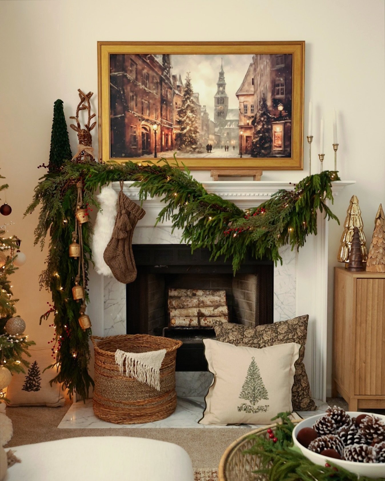 White marble fireplace with a painting of a snowy European street scene, draped with thick cedar garland, knit stockings, and small copper bells.