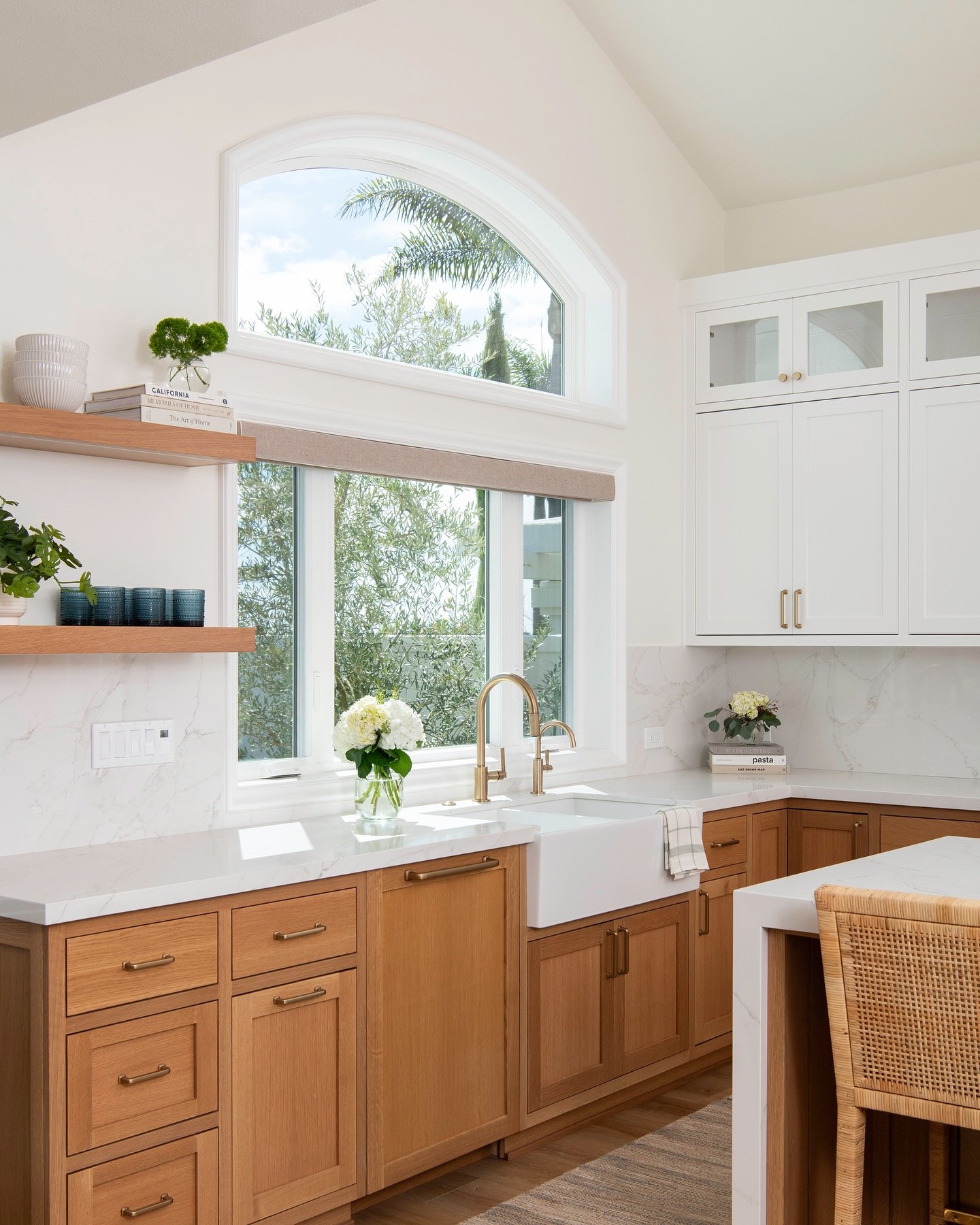 This window treatment uses two stacked windows to capitalize on the kitchen's vaulted ceiling height. The lower rectangular window provides a clean view and direct natural light over the sink. The upper half-circle transom fills the high space, drawing the eye up and enhancing the room's spacious feel. Warm, natural wood cabinets below contrast beautifully with the white upper cabinets, creating a modern coastal vibe. This combination is an excellent strategy for flooding a kitchen with light while adding significant architectural interest.