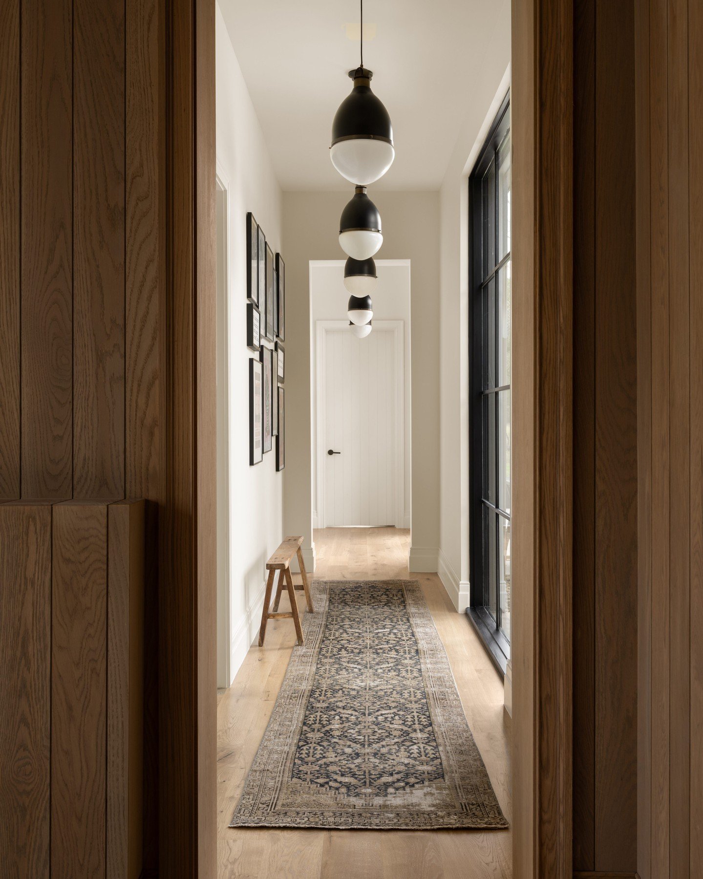 Narrow modern hallway with vertical wood paneling, light wood floor, dark runner rug, and a descending series of five black and white globe pendant lights