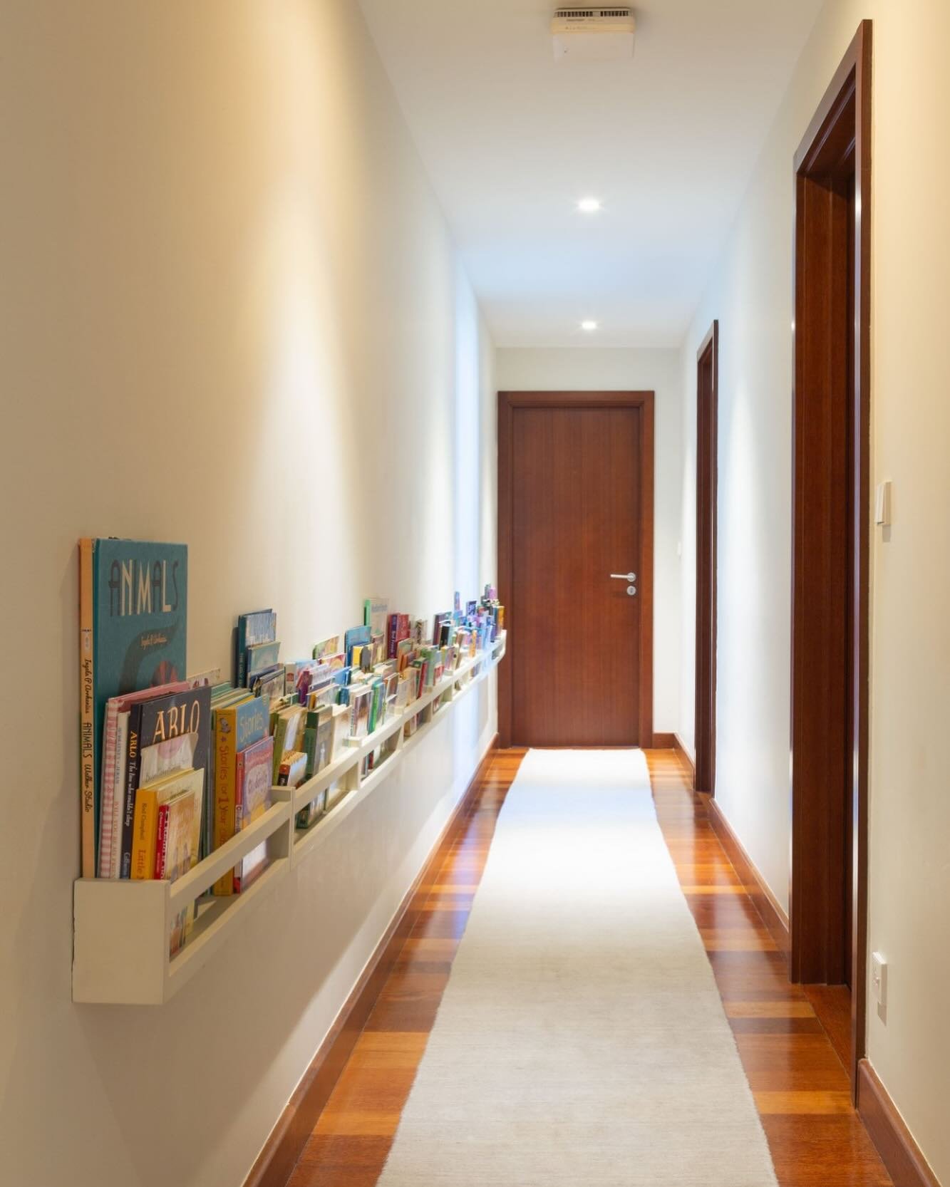 A long, narrow white picture ledge bookshelf mounted low on a hallway wall, displaying the covers of colorful children's books facing outward.